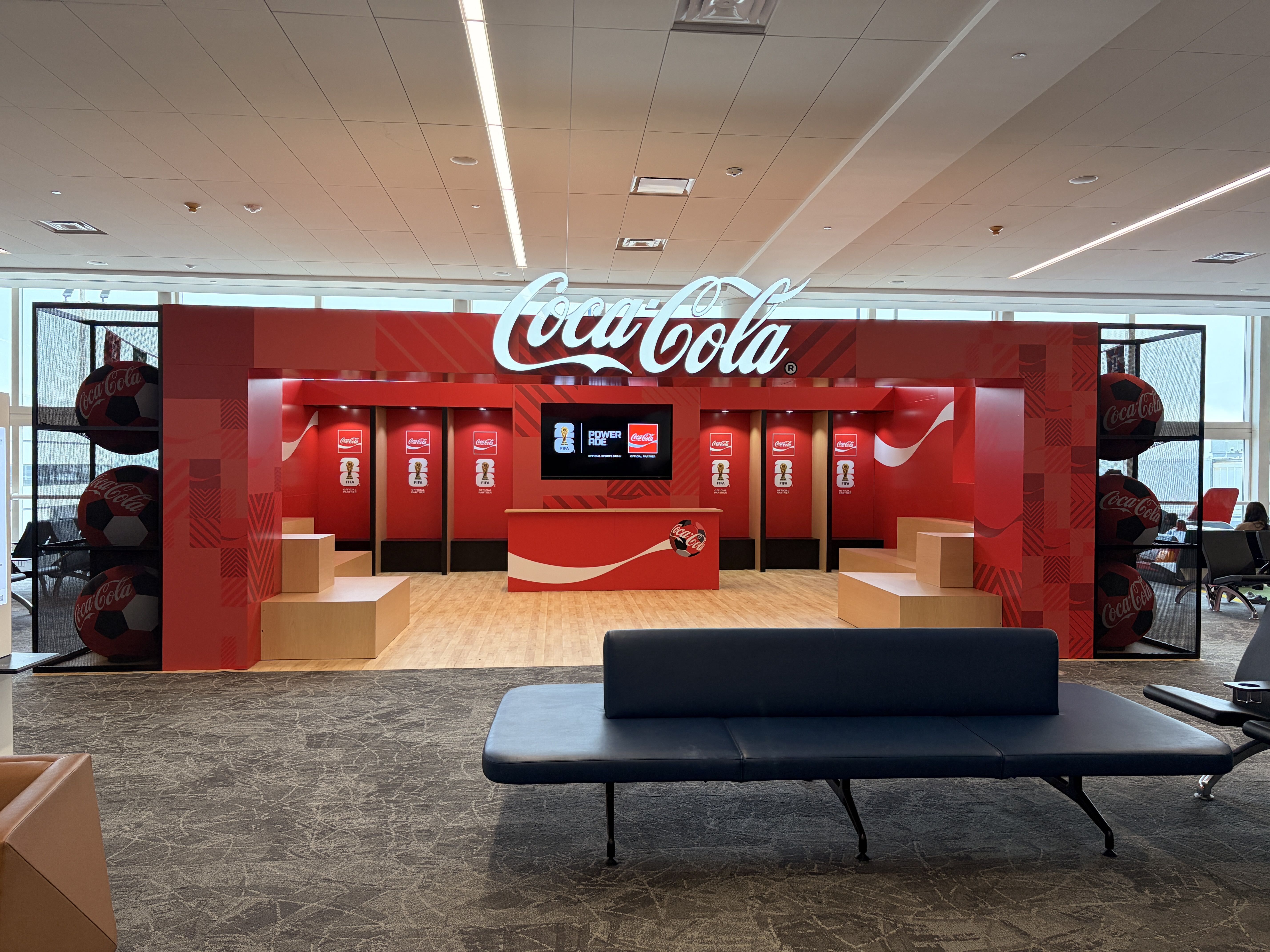 Interior Coca-Cola themed display area with a large white Coca-Cola script sign atop red cabinetry, surrounded by red panels, screens, and oversized Coca-Cola soccer ball props near glass walls.