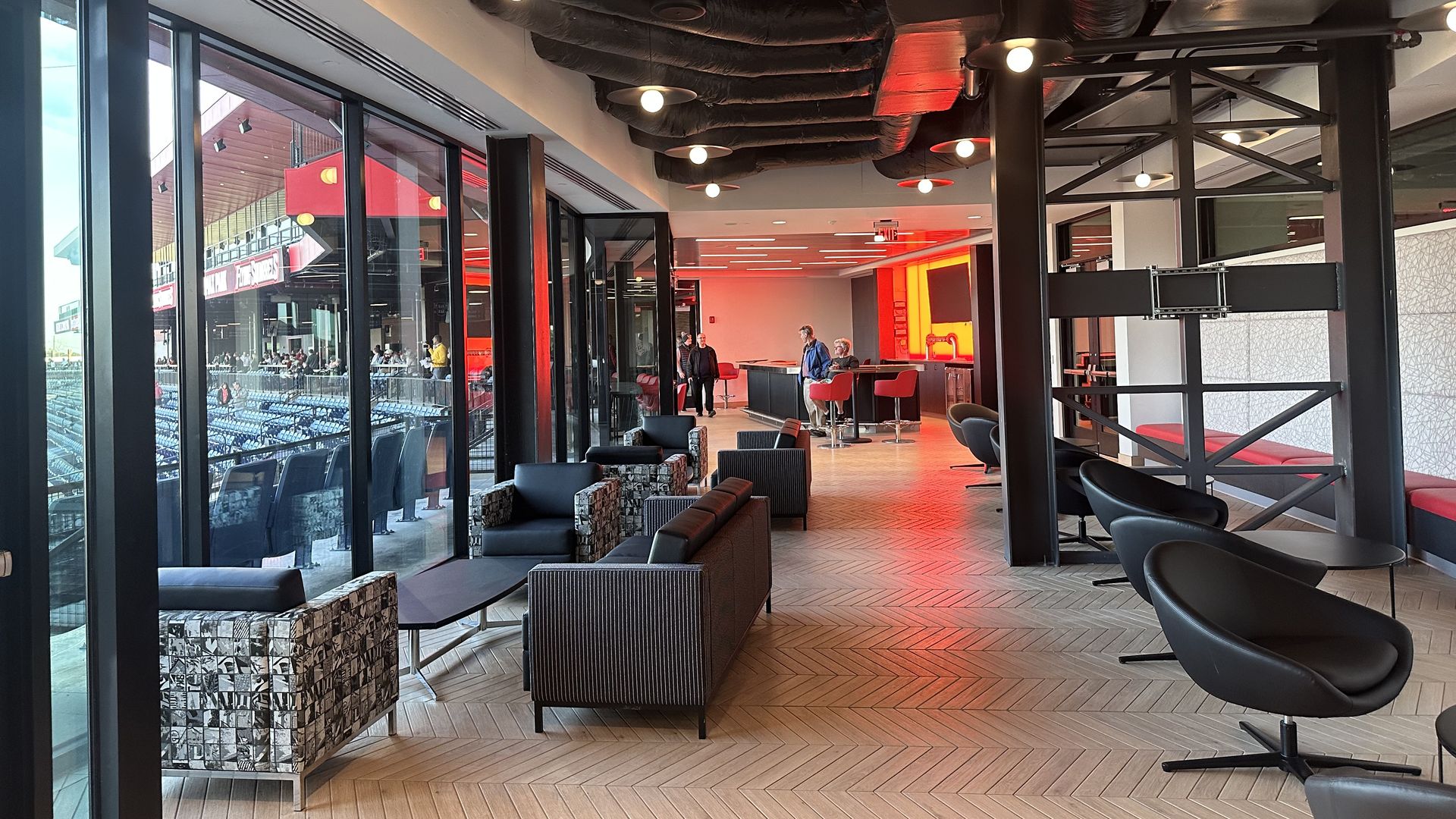 Modern stadium lounge with glass walls facing empty seating. Exposed black ductwork ceiling, round lights, and patterned chairs with black sofas; red-lit bar at the far end.