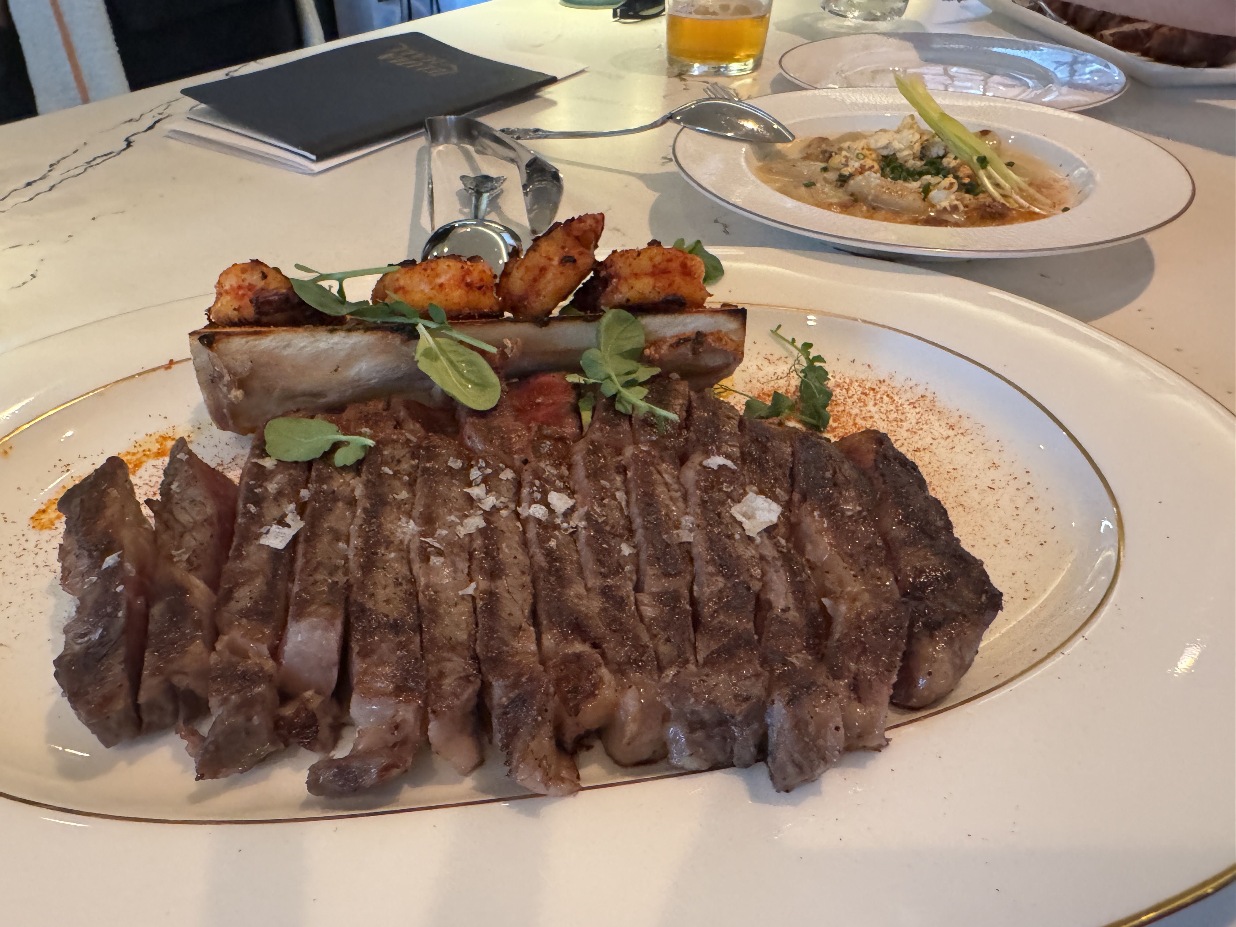Sliced grilled steak garnished with coarse salt and greens, served on an oval plate with a bone topped with shrimp. A bowl of creamy soup and a glass of beer are in the background.