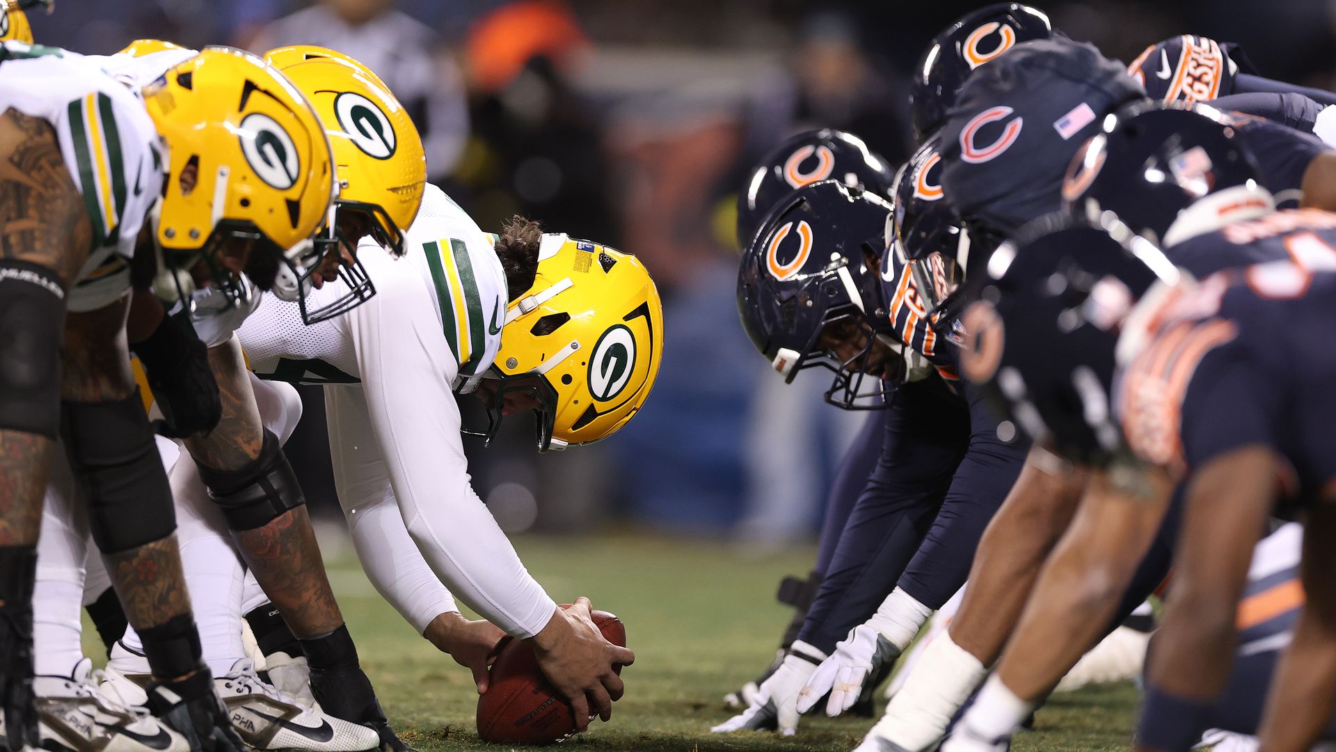 Football players from the Green Bay Packers in yellow helmets and white uniforms facing off against Chicago Bears players in navy helmets and dark uniforms, lined up before a play.