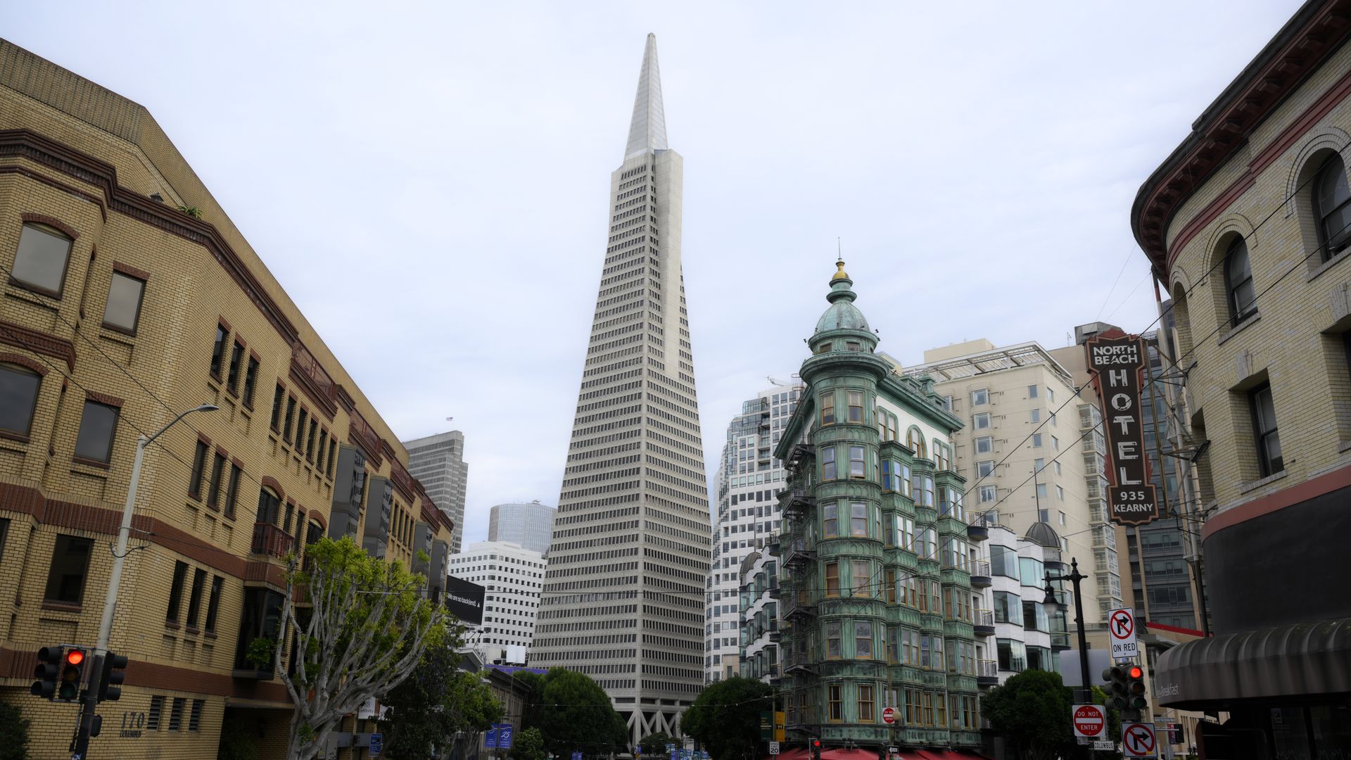 City street with the tall, pointed Transamerica Pyramid building in the center, a historic green copper building with red umbrellas on the right, and a yellow brick building on the left under a cloudy sky.