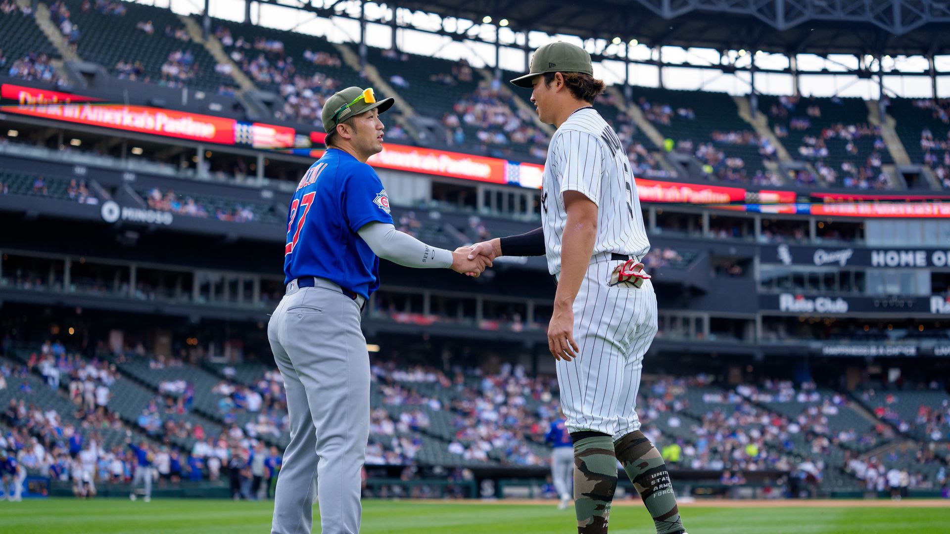 Two baseball players shake hands on the field amid a stadium crowd: one in a blue jersey with a green cap, the other in a white pinstripe uniform. Grass and stands visible in the background.