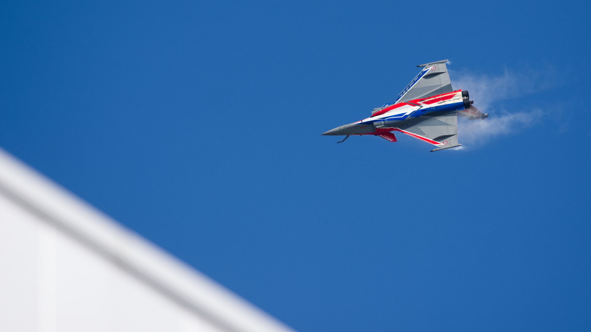 A fighter jet banks hard through a clear blue sky. Vapor shoots off its tail. The roof of a nearby building is seen in the corner.