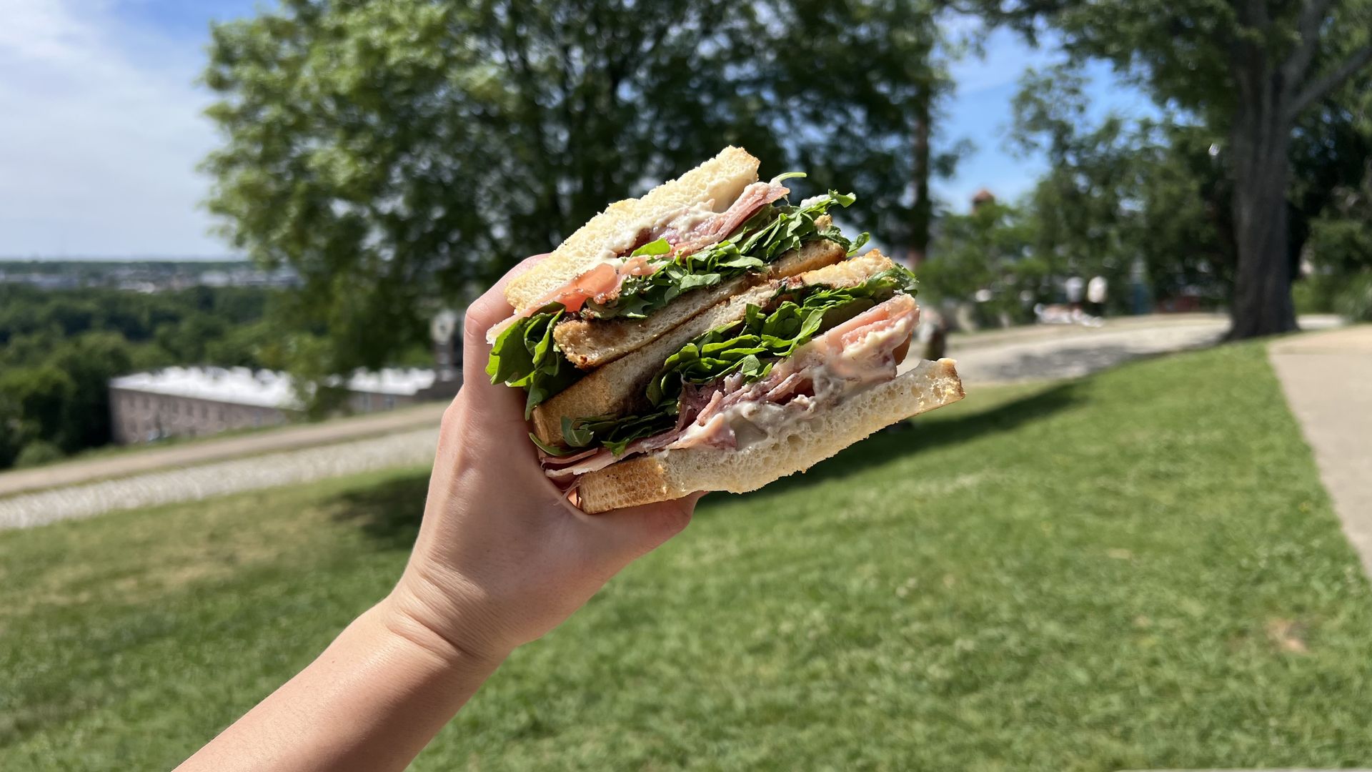 a giant sandwich being held in the air by a hand. In the background is a park with greenery