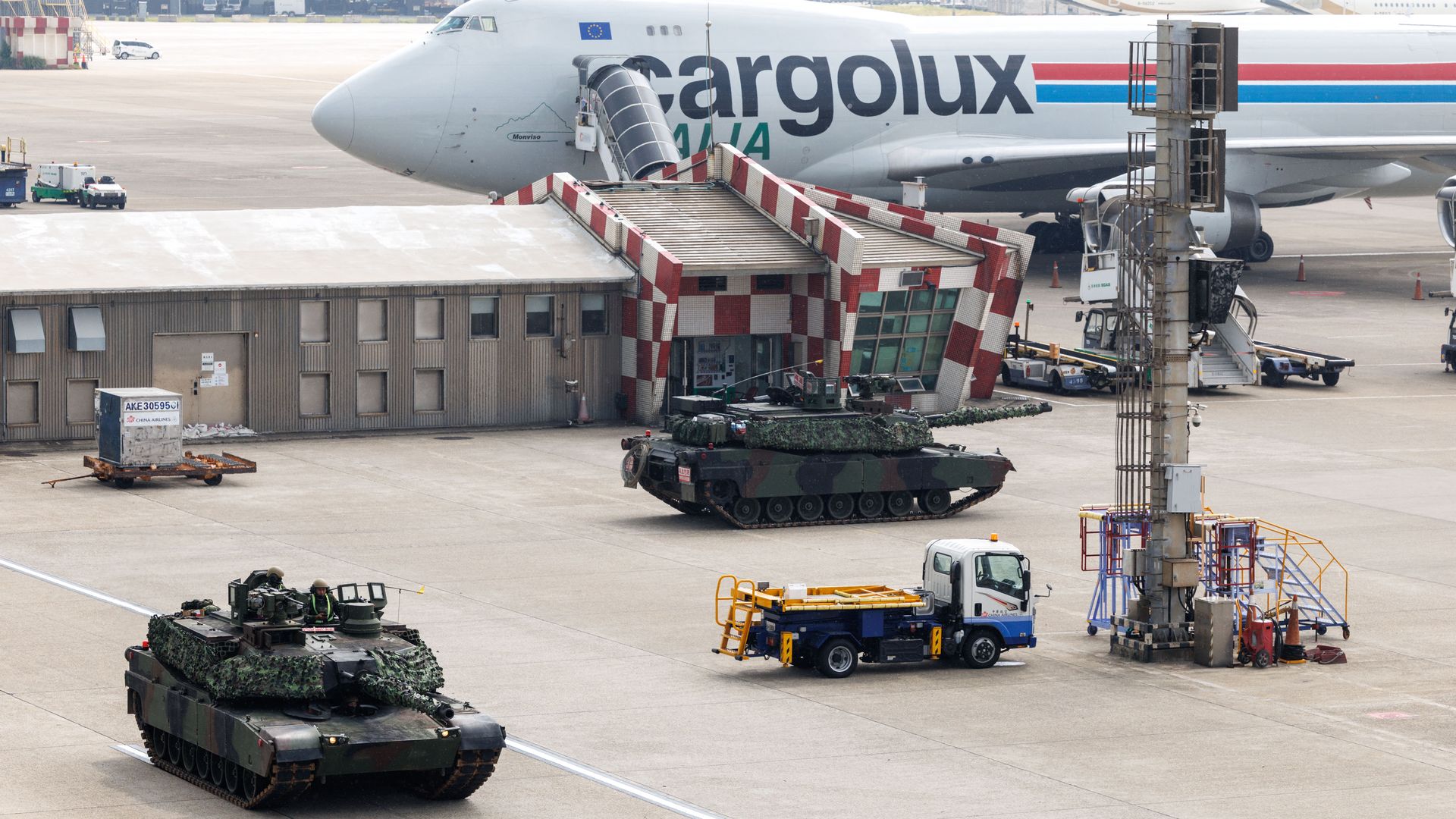A wide view of a Cargolux cargo aircraft at an airport gate. A jet bridge attaches to the door. Camouflaged tanks and ground vehicles are on the tarmac.