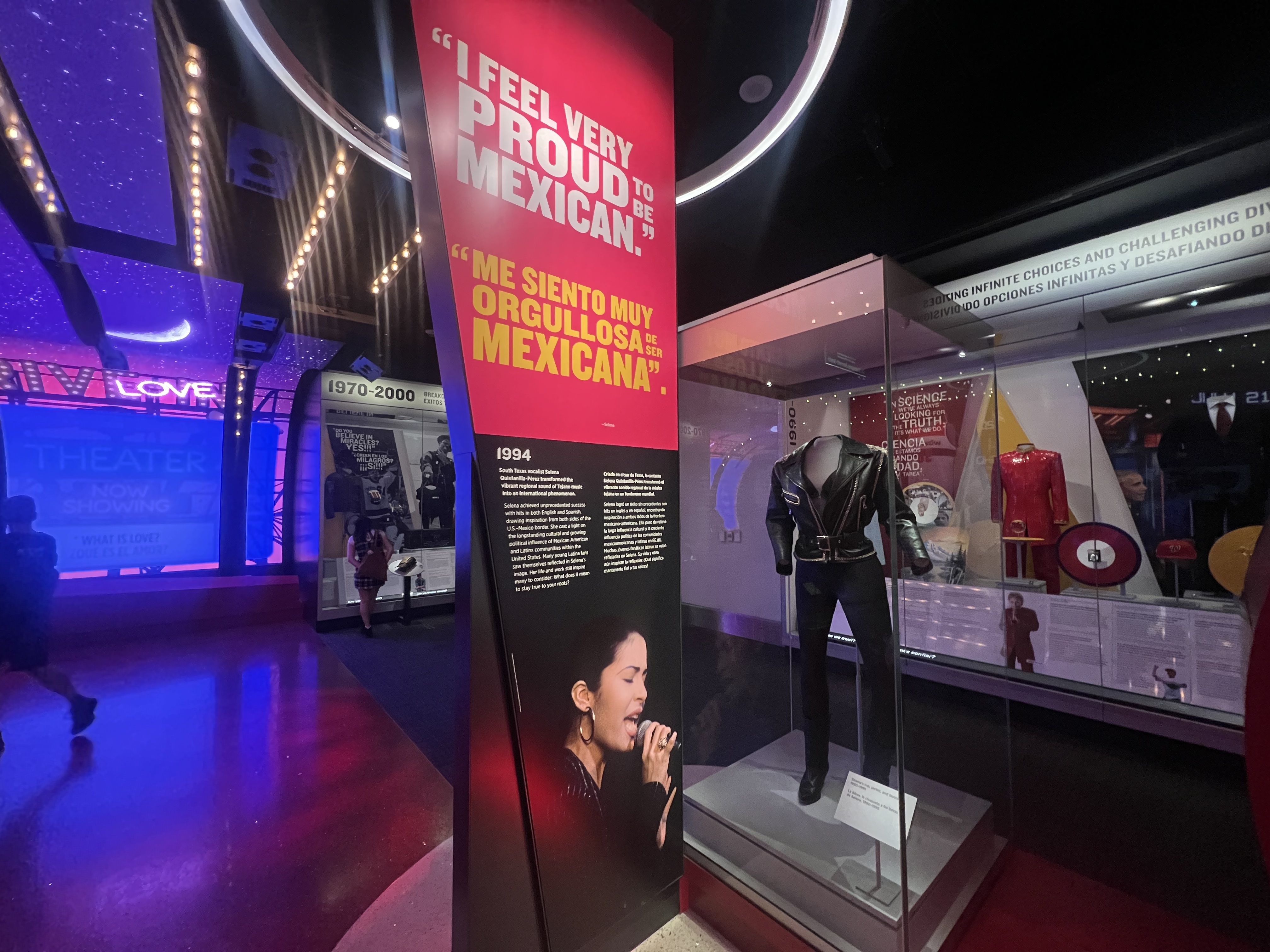 Museum exhibit featuring a leather jacket and microphone from 1994 with a pink sign quoting "I feel very proud to be Mexican." in English and Spanish. Purple and blue lighting.