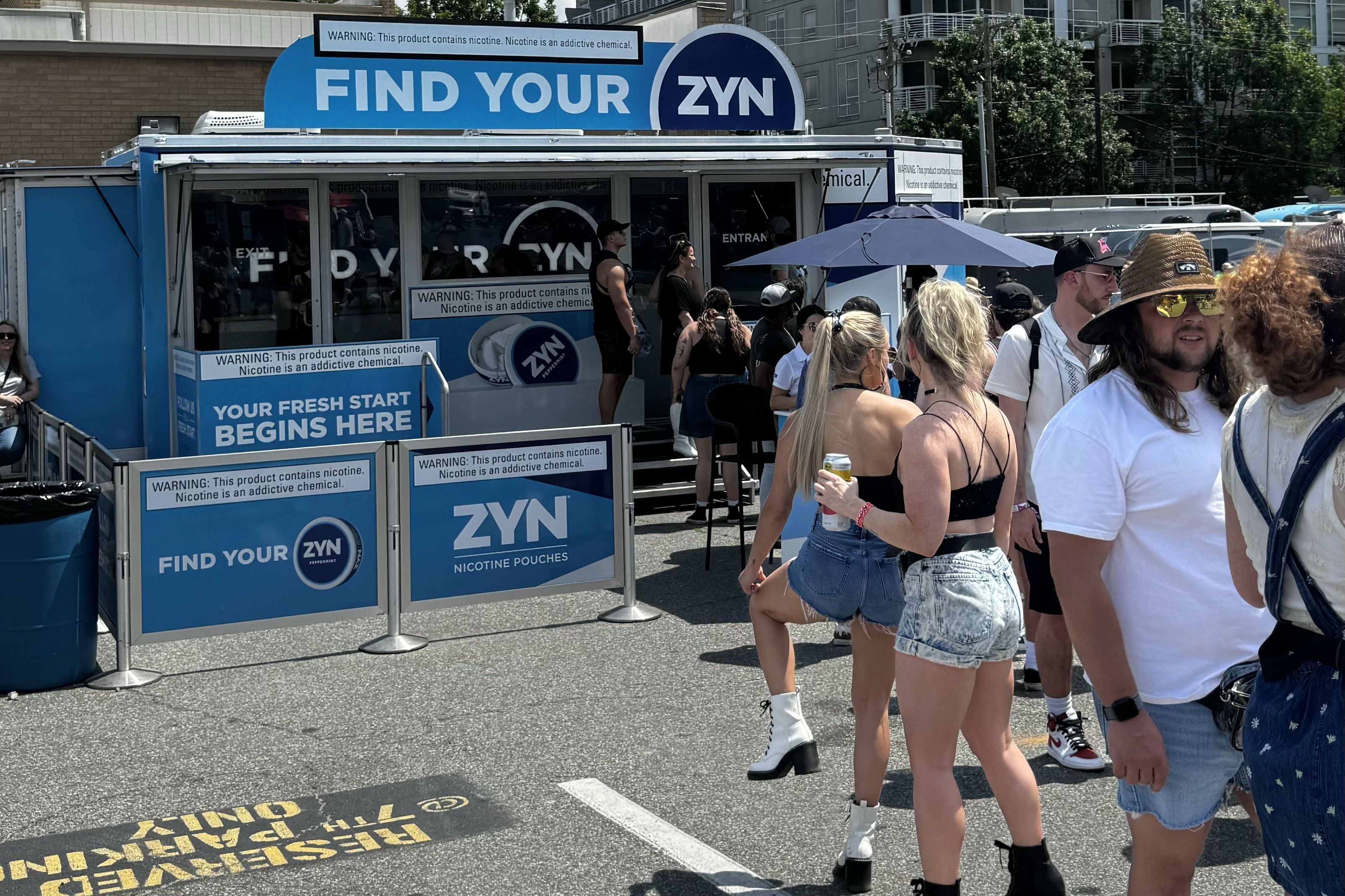 Blue promotional trailer reading "FIND YOUR ZYN" advertising nicotine pouches; a crowd lines up outside on a sunny street with umbrellas and city buildings in the background.
