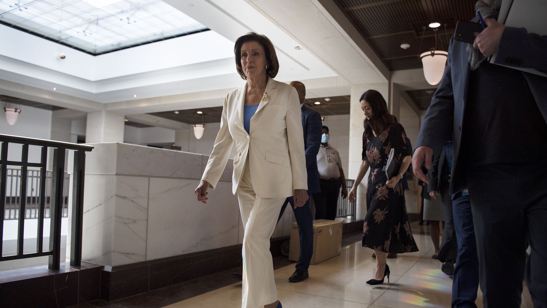 Photo of Nancy Pelosi in a white suit and blue heels walking in the Capitol hallway