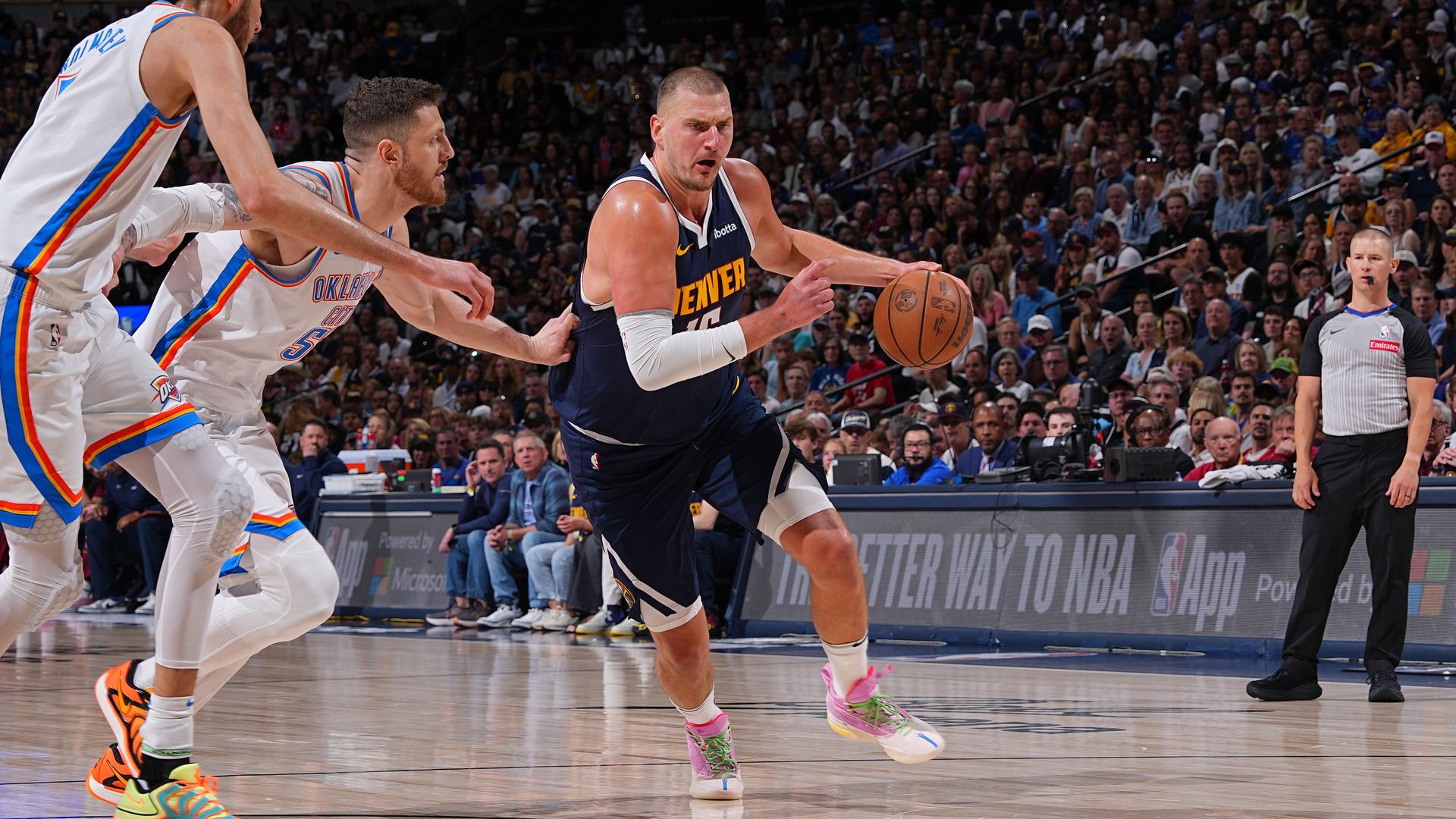 Denver Nuggets player dribbling basketball fiercely guarded by two Oklahoma City Thunder players in white uniforms during a packed NBA game with referee and crowd visible.