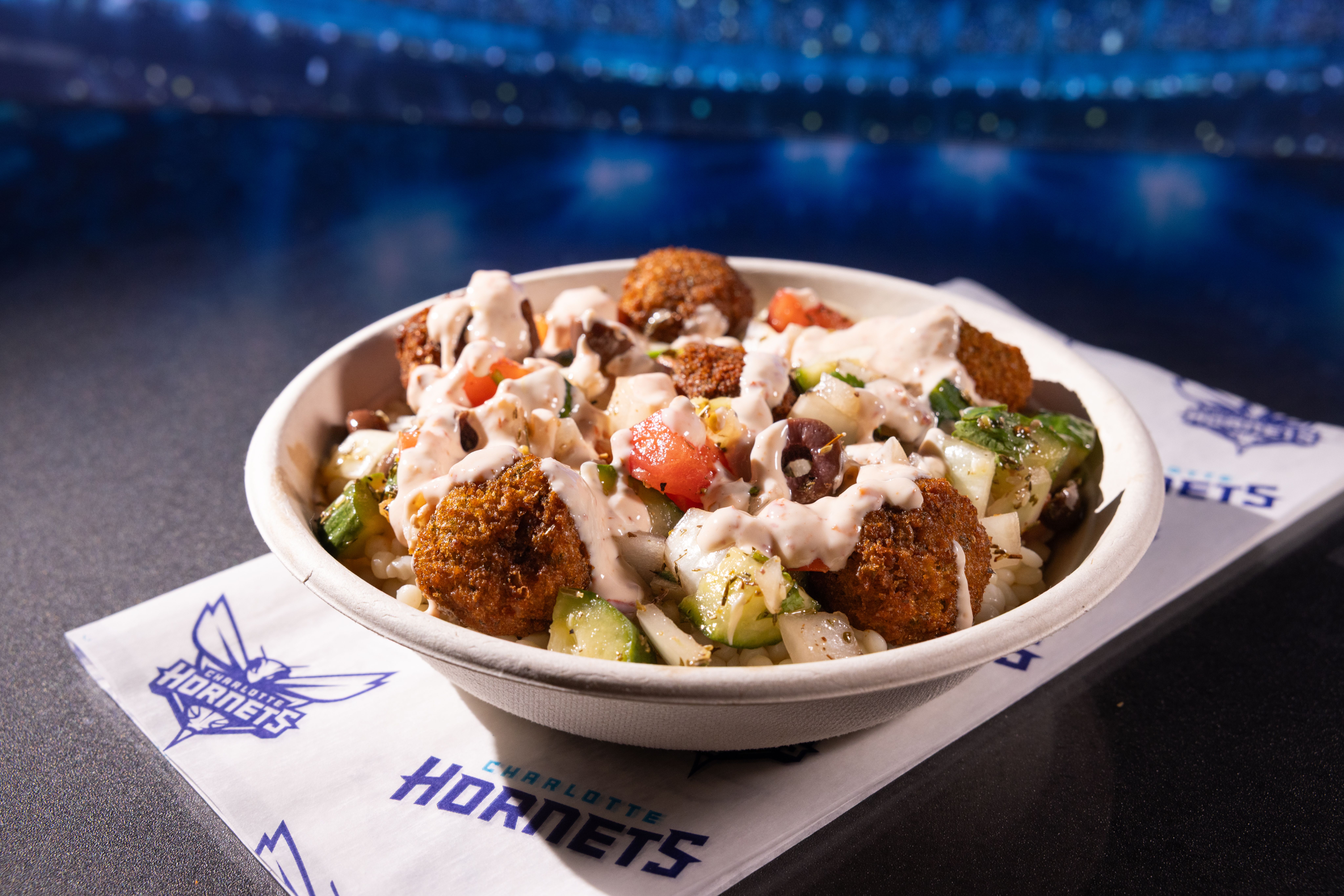 Bowl of falafel balls with chopped cucumbers, tomatoes, olives, onions, and creamy dressing on top, placed on a Charlotte Hornets branded napkin on a dark surface.