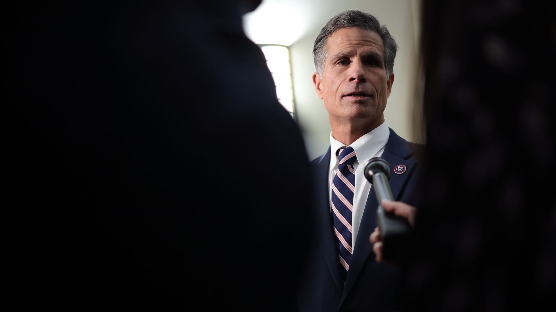 A photo of a man being interviewed between the silhouettes of two onlookers in the foreground. 