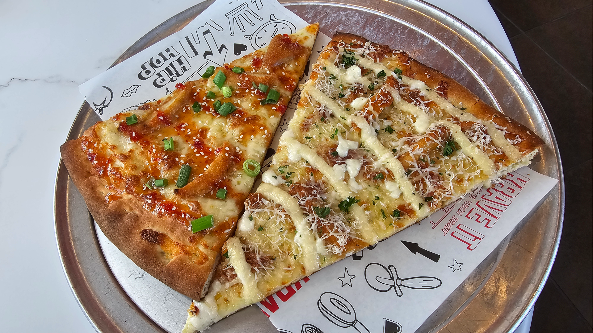 Two rectangular pizza slices on a metal tray with printed parchment. Left slice has red sauce, cheese, green onions and sesame seeds; right slice has white sauce drizzle, cheese shreds, herbs.