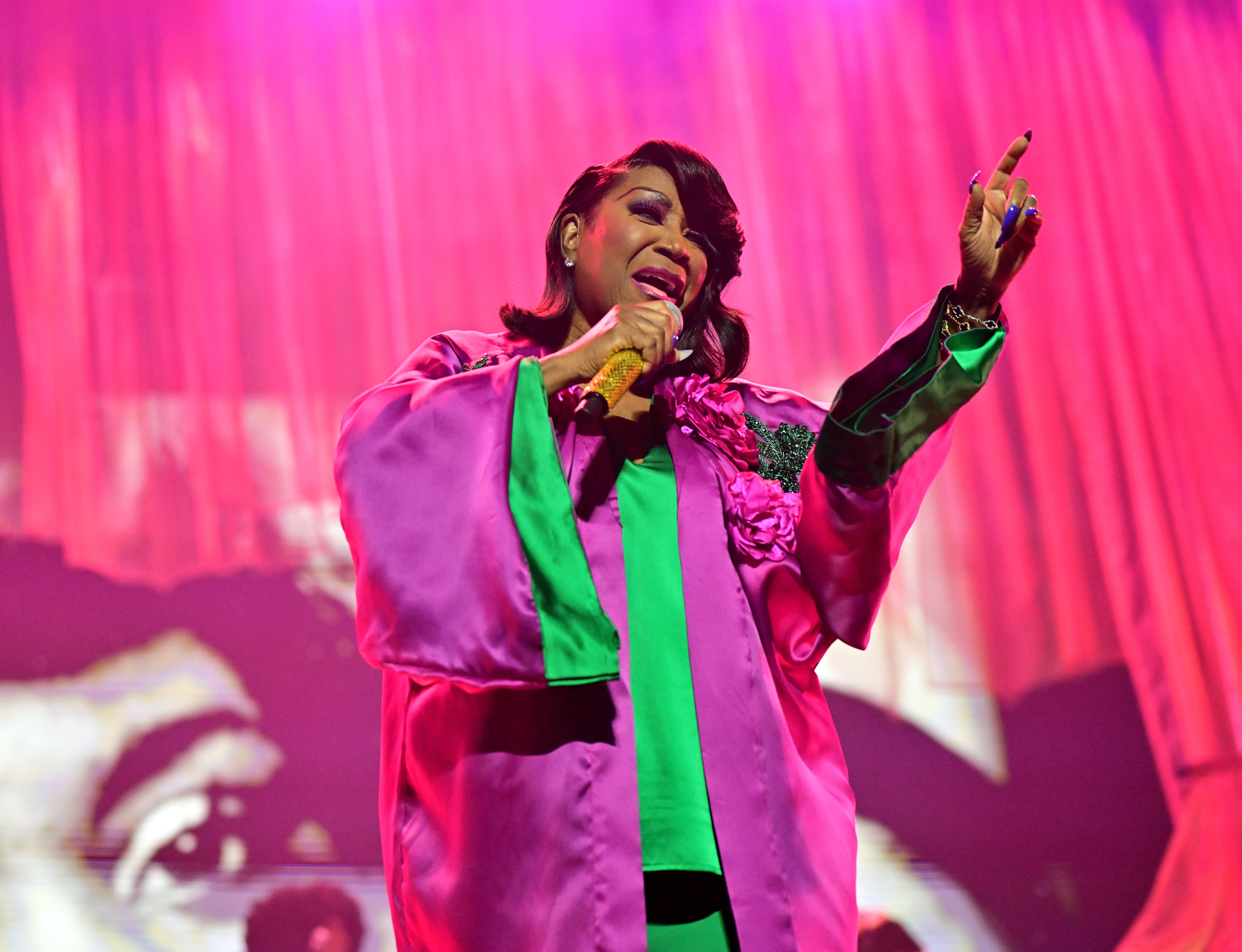 A singer in a bright pink and green outfit passionately performs on stage with a pink backdrop, holding a glittering gold microphone and gesturing with one hand raised.