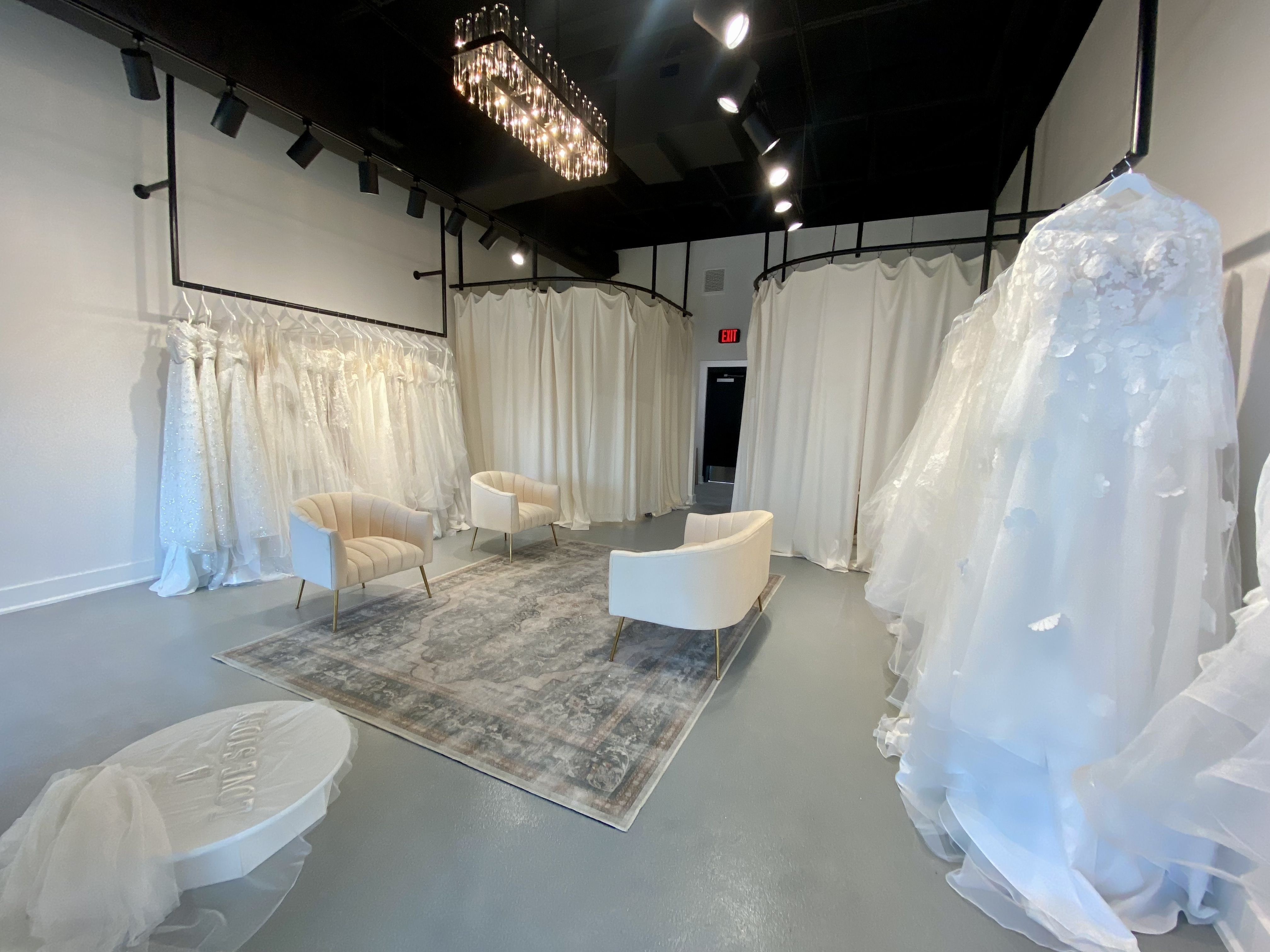 Bright bridal boutique with white wedding dresses hanging on black racks, three cream chairs around a patterned rug, white curtains, and a modern chandelier above.