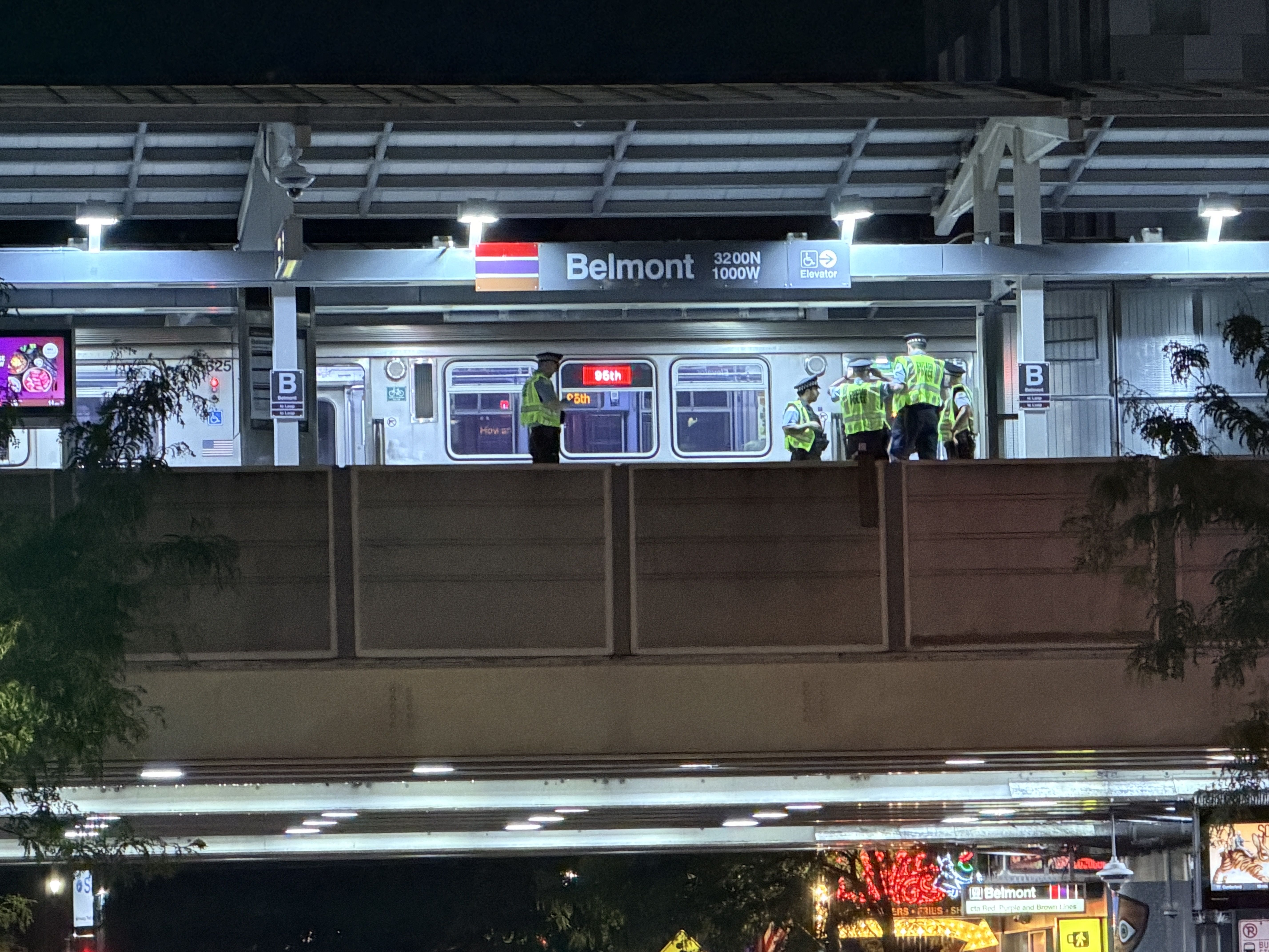 Night scene at Belmont station. A silver subway car on an elevated platform with several workers in bright yellow vests near the doors. Overhead beams and a blue Belmont sign with a flag.