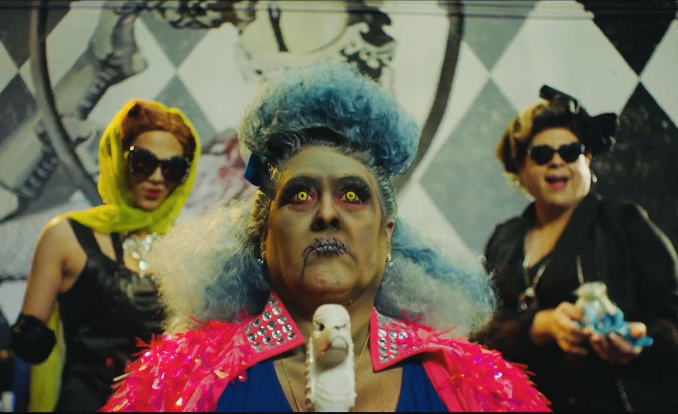 A dramatically styled person in a bright pink sequined jacket with blue hair and yellow eyes stands in the center, holding a white bird. Their face is heavily painted, with dark eye makeup and stitched lips. Two other individuals are in the background, both wearing sunglasses and elaborate costumes.