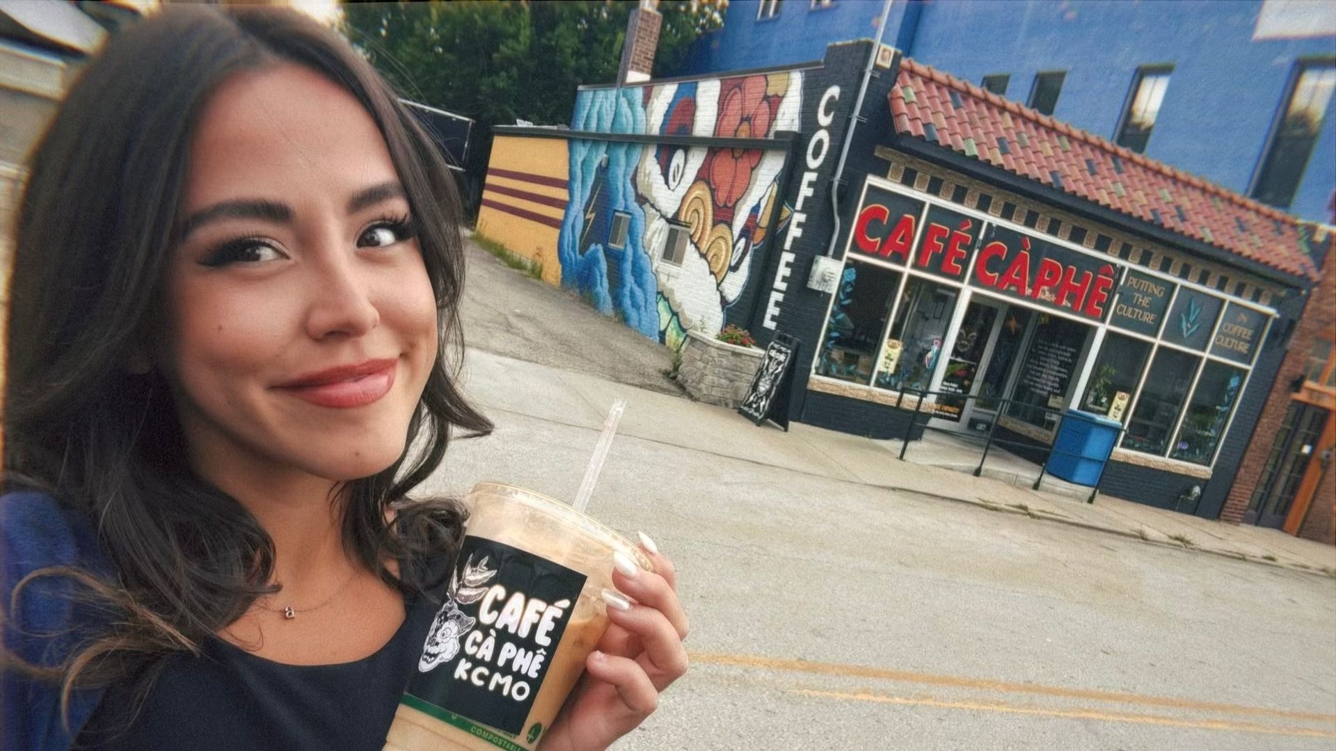 A young woman with dark hair smiles holding an iced coffee outside a colorful building with red "CAFÉ CÀ PHÊ" sign and mural, wearing a navy top and blue jacket.