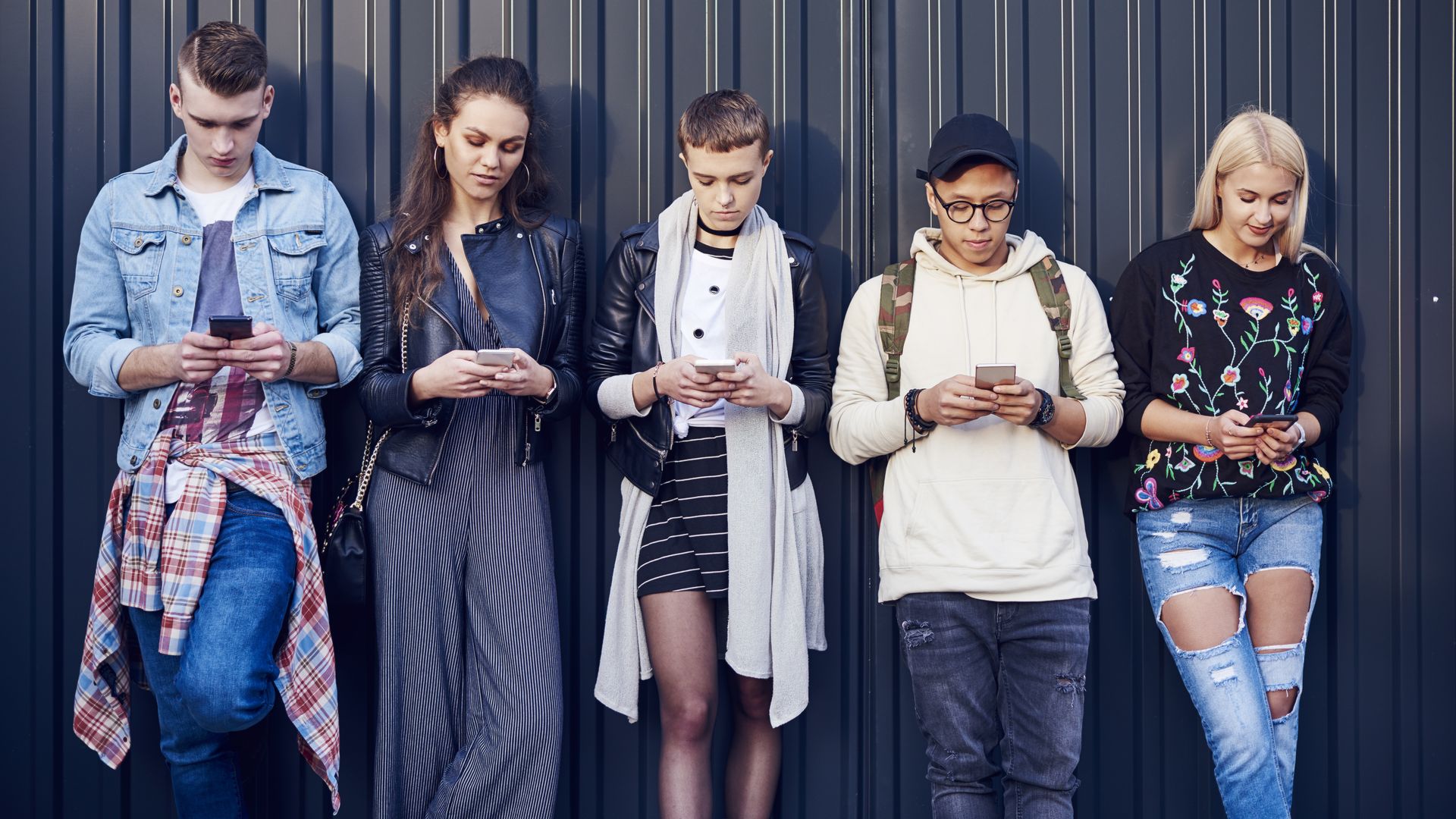 In this image, five fashionably dressed young adults look at their phones while standing in a line against a wall.