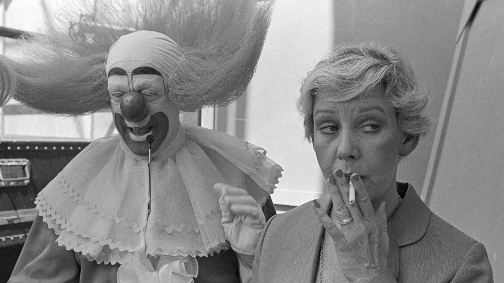Photo of a clown next to a woman smoking a cigarette. 