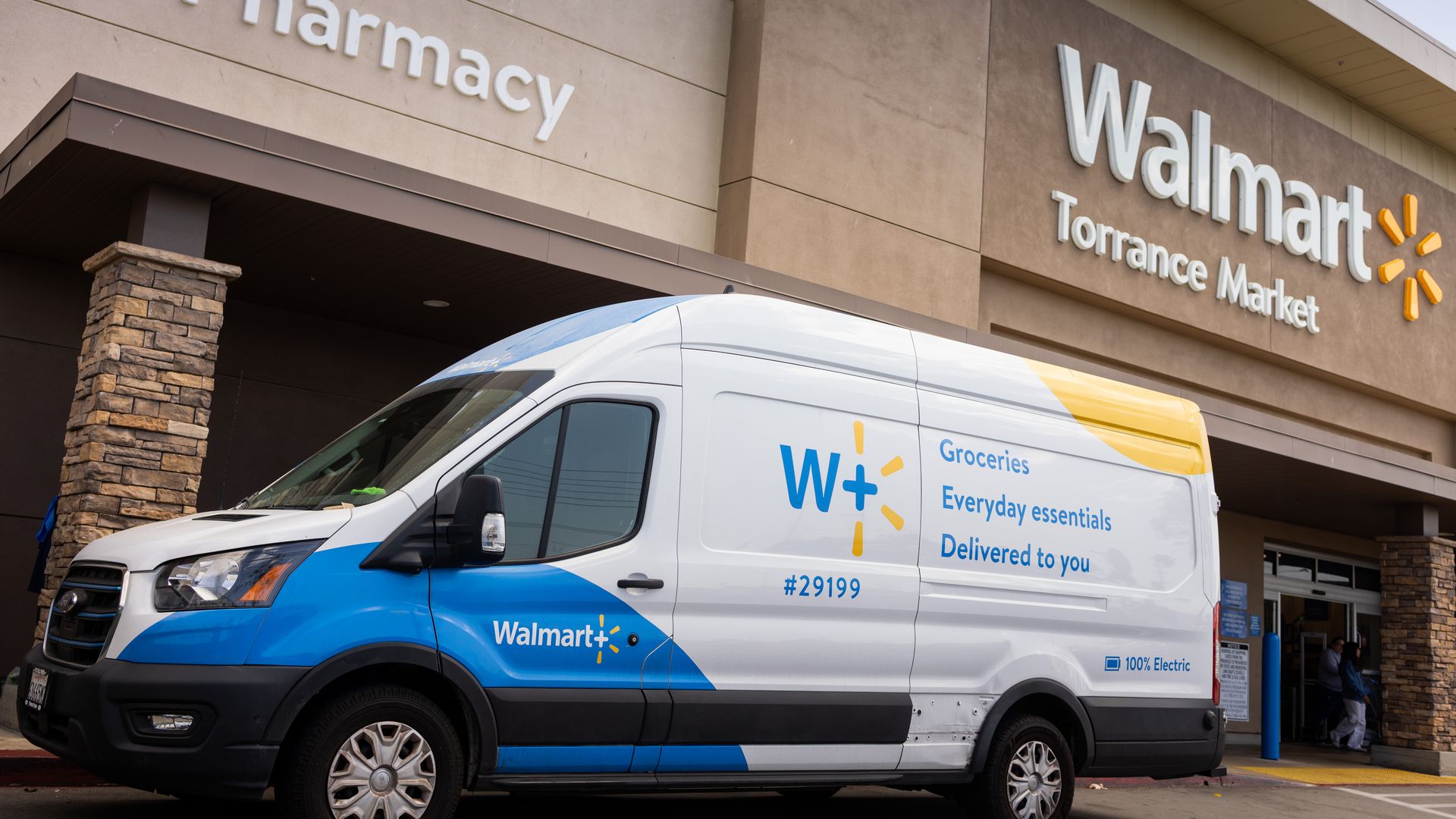 Exterior of a Walmart store with a large white Walmart sign and the word "pharmacy" on a beige wall; a white and blue Walmart delivery van is parked in front.