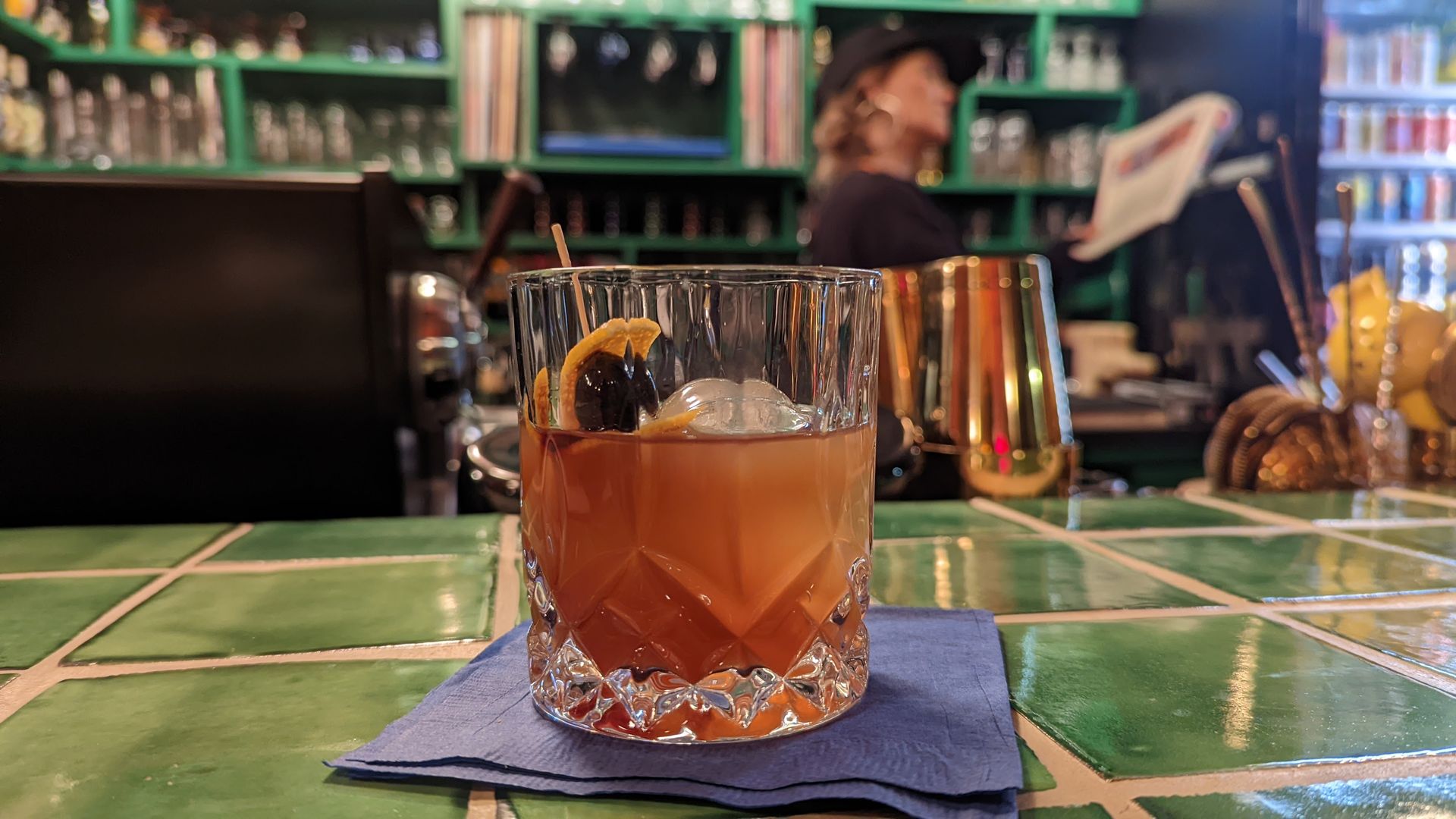 A cocktail sits on a blue napkin and contains a round ice cube and a Luxardo cherry and orange peel.