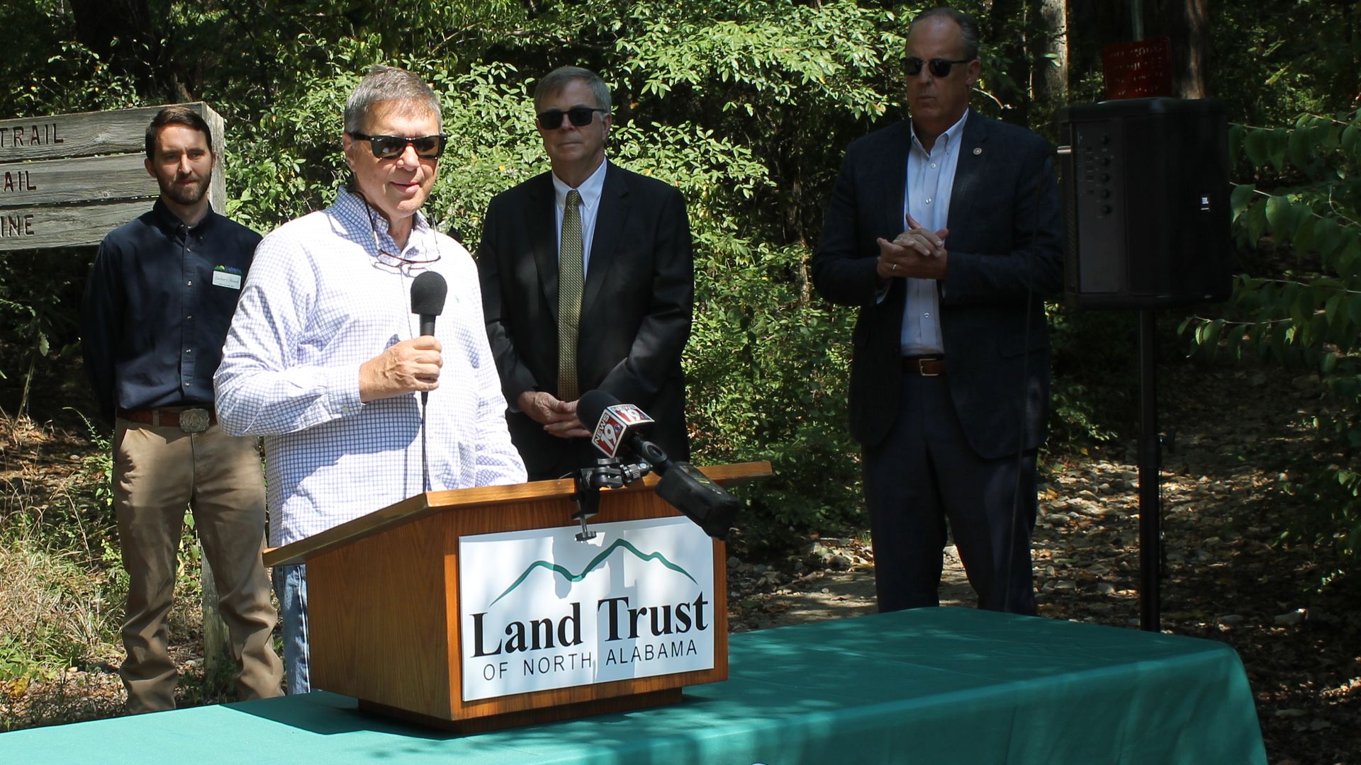 Outdoor event with six people standing near a green table covered with a "Land Trust of North Alabama" cloth; one man at a podium with microphone speaks, a wooded trail sign is in the background.