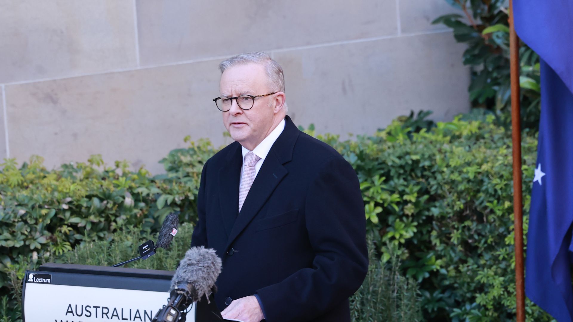 Man in black coat and pink tie, Australian Prime Minister Anthony Albanese, speaking at Australian War Memorial lectern during Last Post Ceremony with Australian flag and greenery in background.