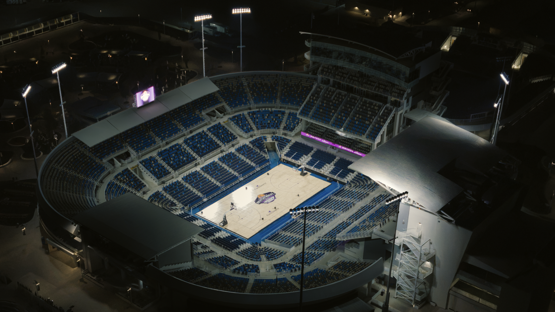 Nighttime aerial view of a circular basketball arena with blue seats surrounding a lit hardwood court. Tall floodlights, a purple LED banner, and surrounding dark concourses.