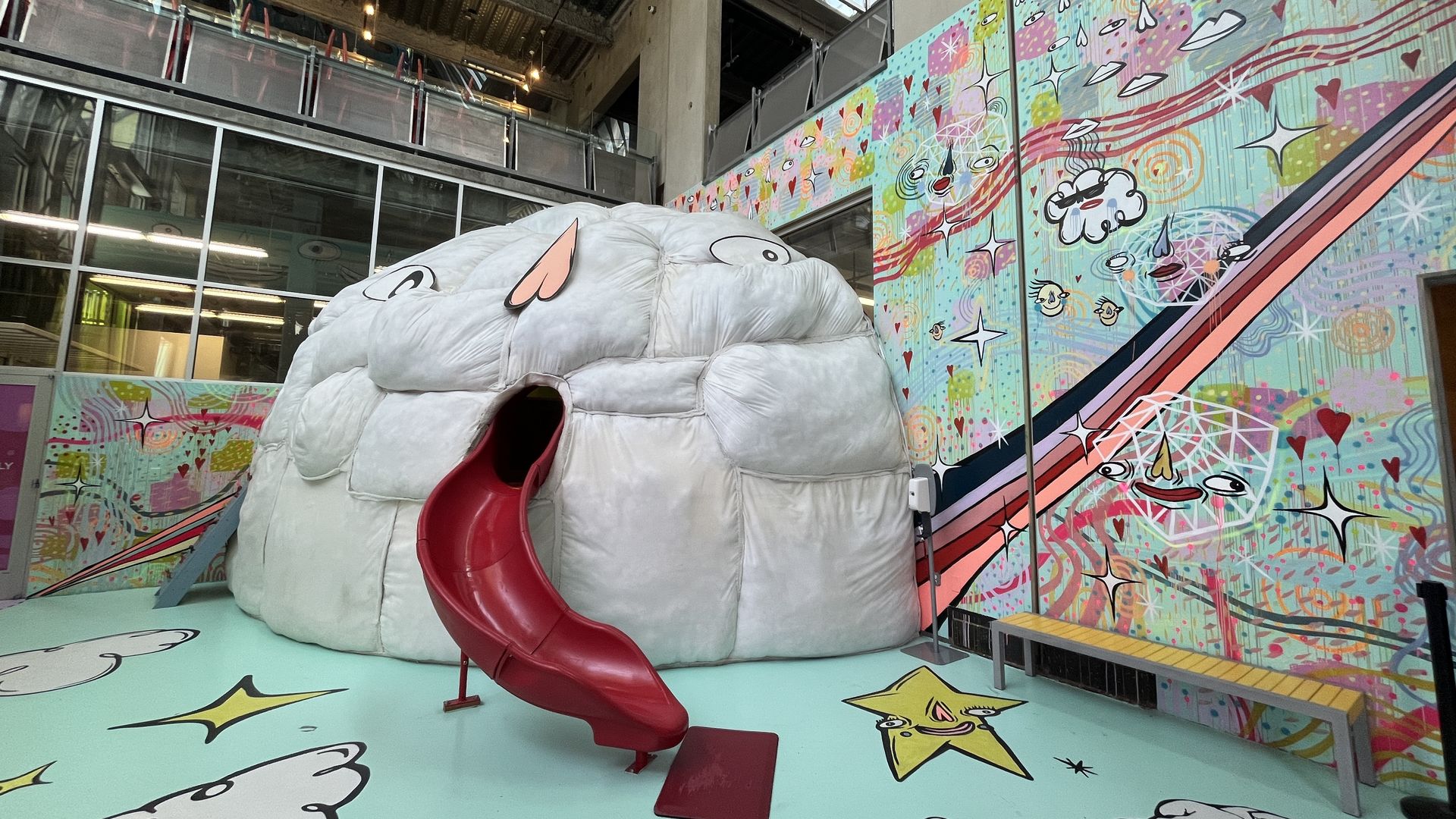 A slide comes out of a cloud character's mouth at a children's museum with rainbow-colored cartoon murals on the wall.