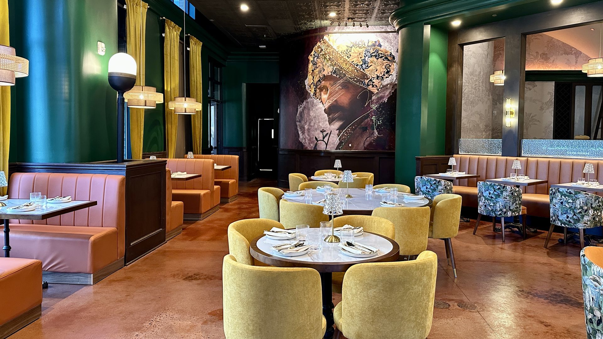 Zamindar's. Chic dining room with emerald green walls, yellow curtains, and orange banquettes. Round tables set with glassware and candles; a large mural at the far wall and decorative lamps along the space.
