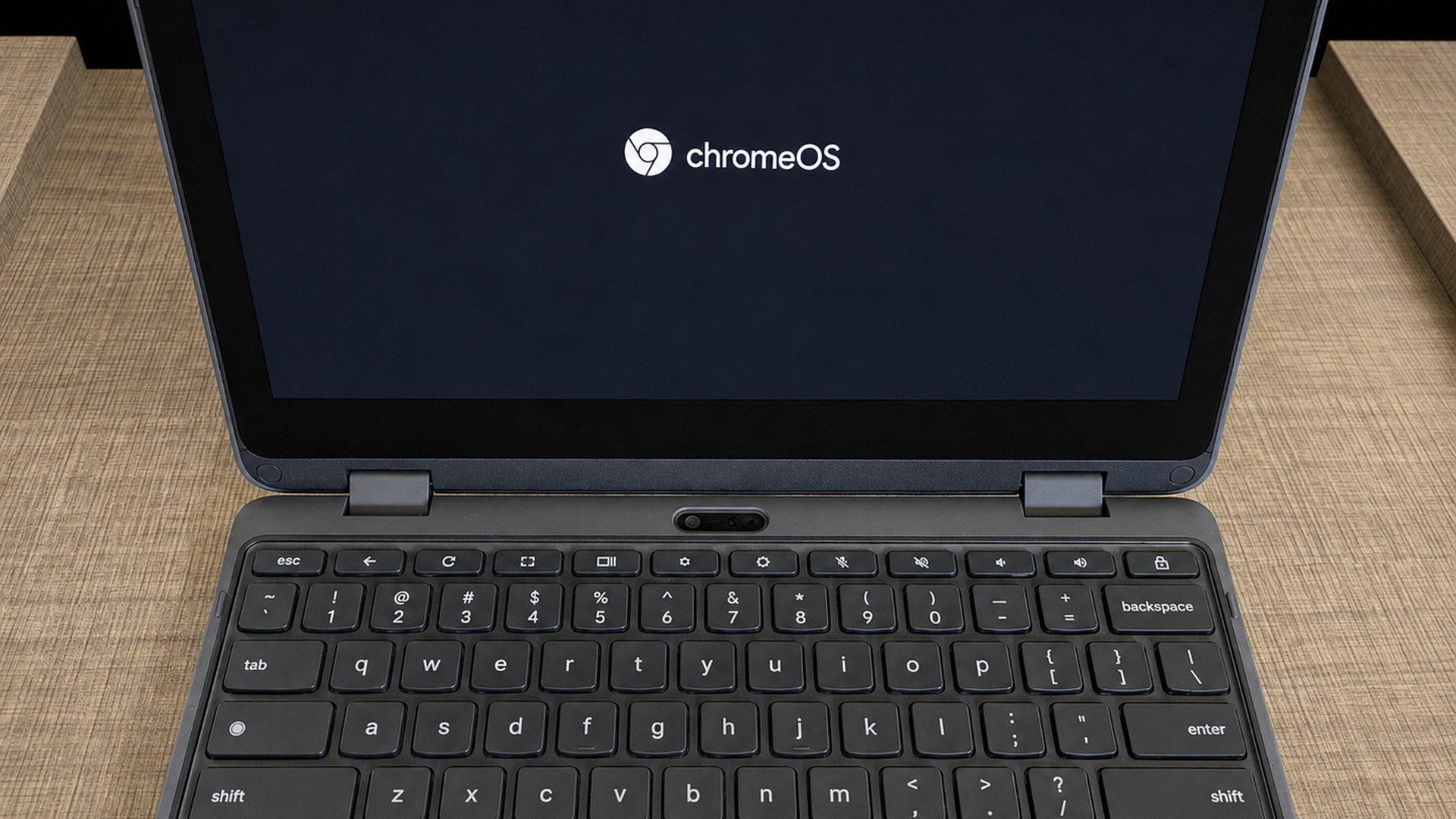 Open laptop on a wooden desk, the dark screen shows the ChromeOS logo. A black keyboard is fully visible with hinges and a centered webcam housing above the keyboard.