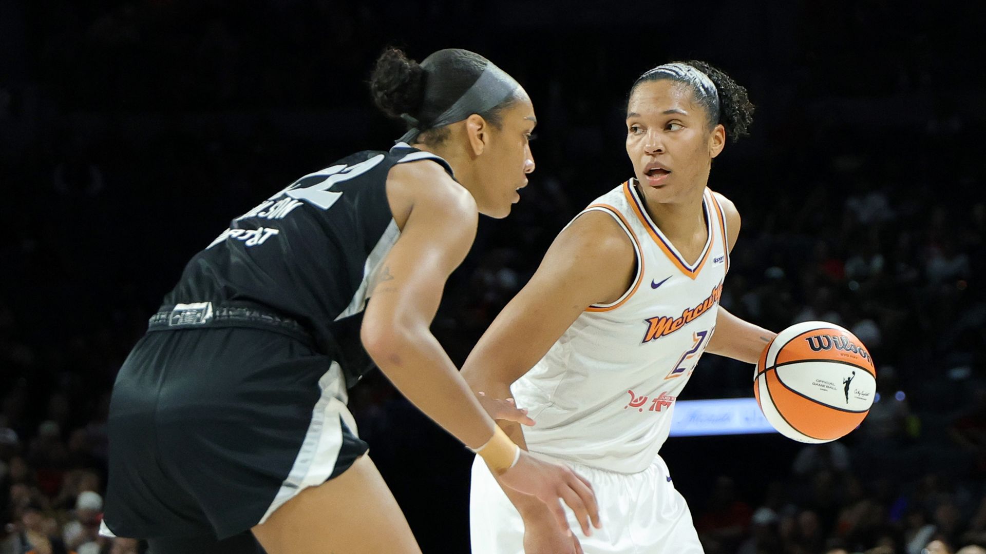 A woman in a white basketball uniform dribbles an orange and white ball against a woman in a black basketball uniform with a crowd in the background.