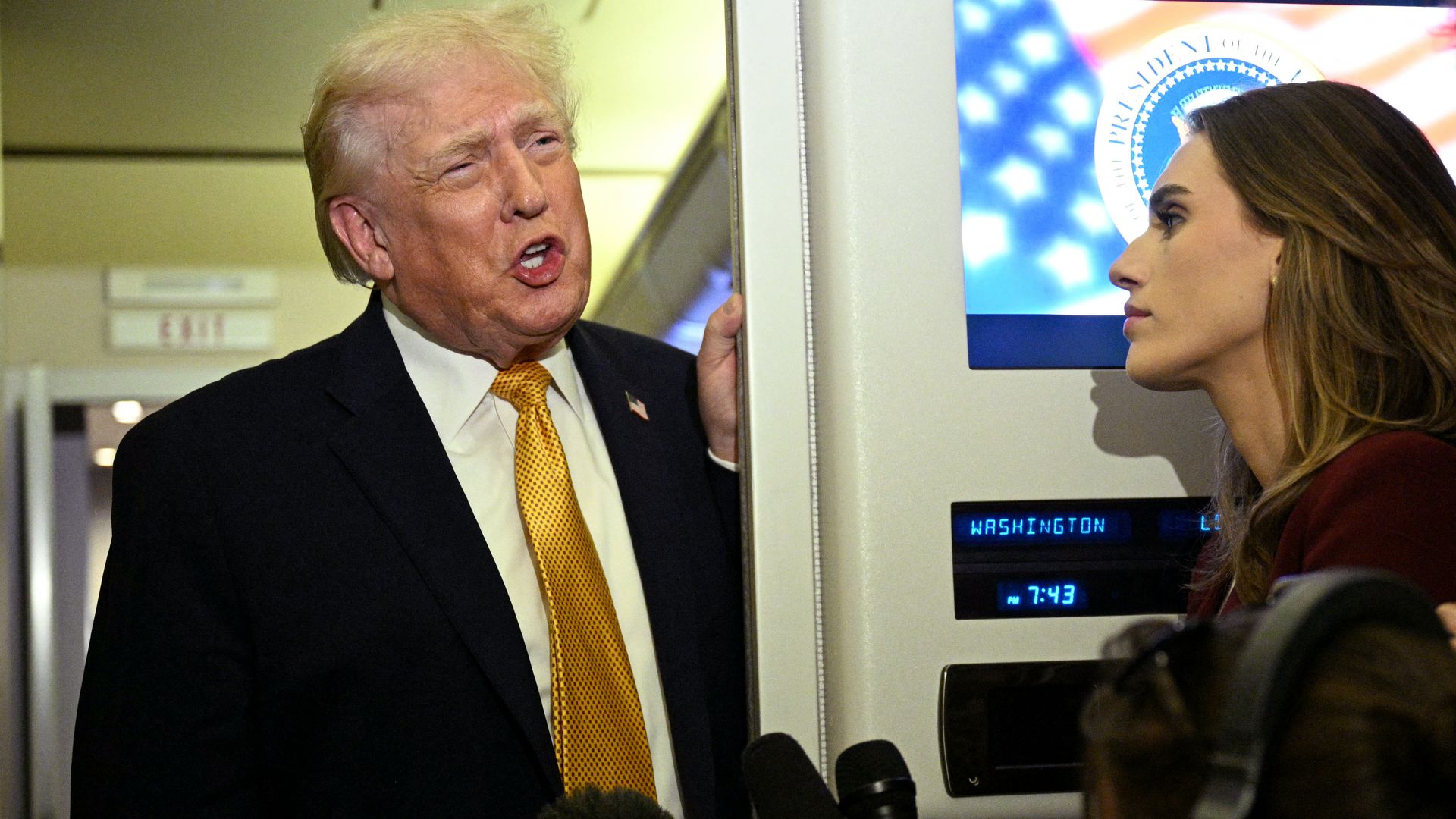 President Donald Trump speaking to reporters, a woman wearing headphones holding microphones, and another man looking at a smartphone inside an aircraft with a screen displaying the presidential seal and US flag.