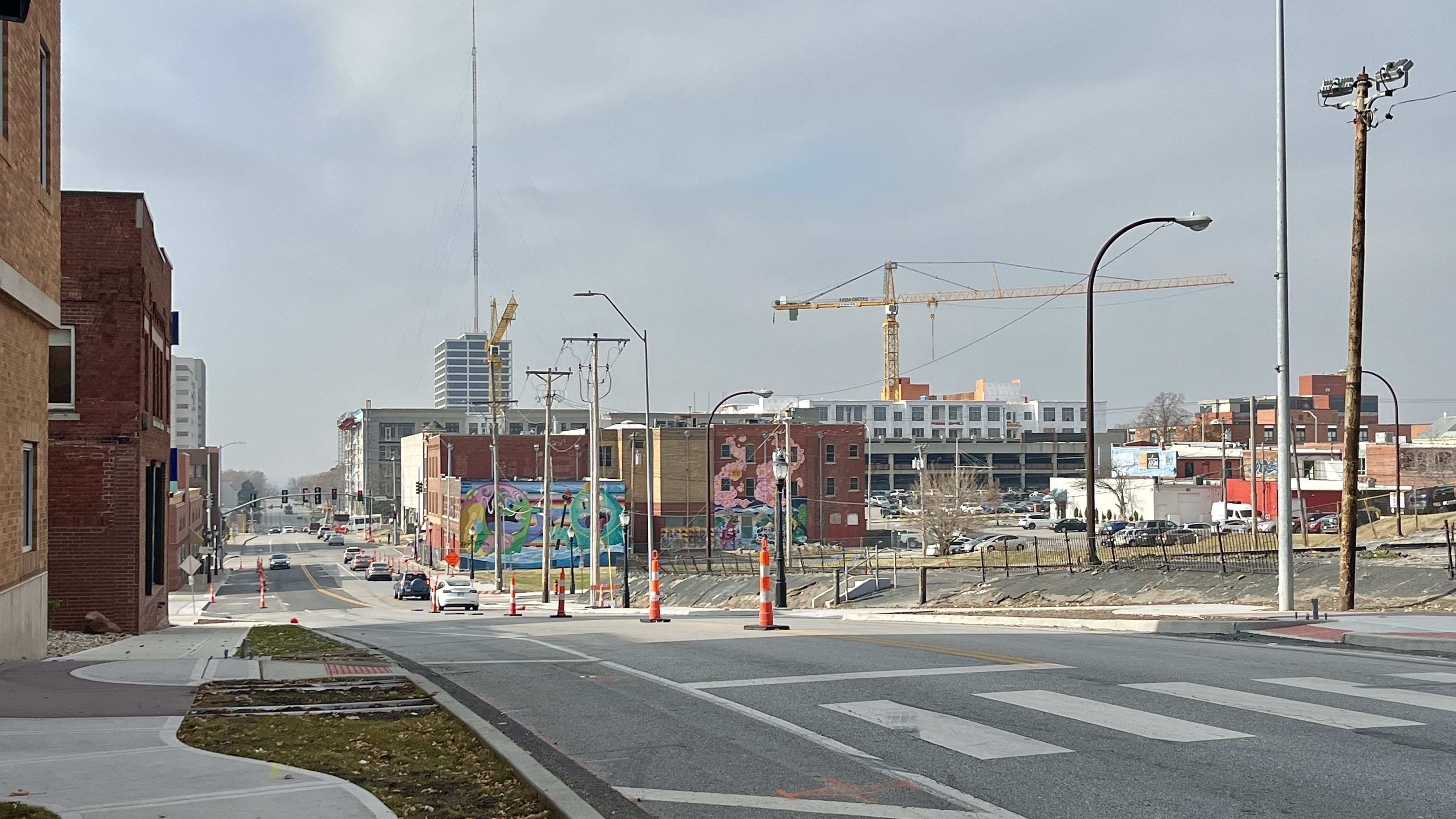 City street with light traffic, orange construction cones, buildings with murals, and cranes in the background under a cloudy sky.