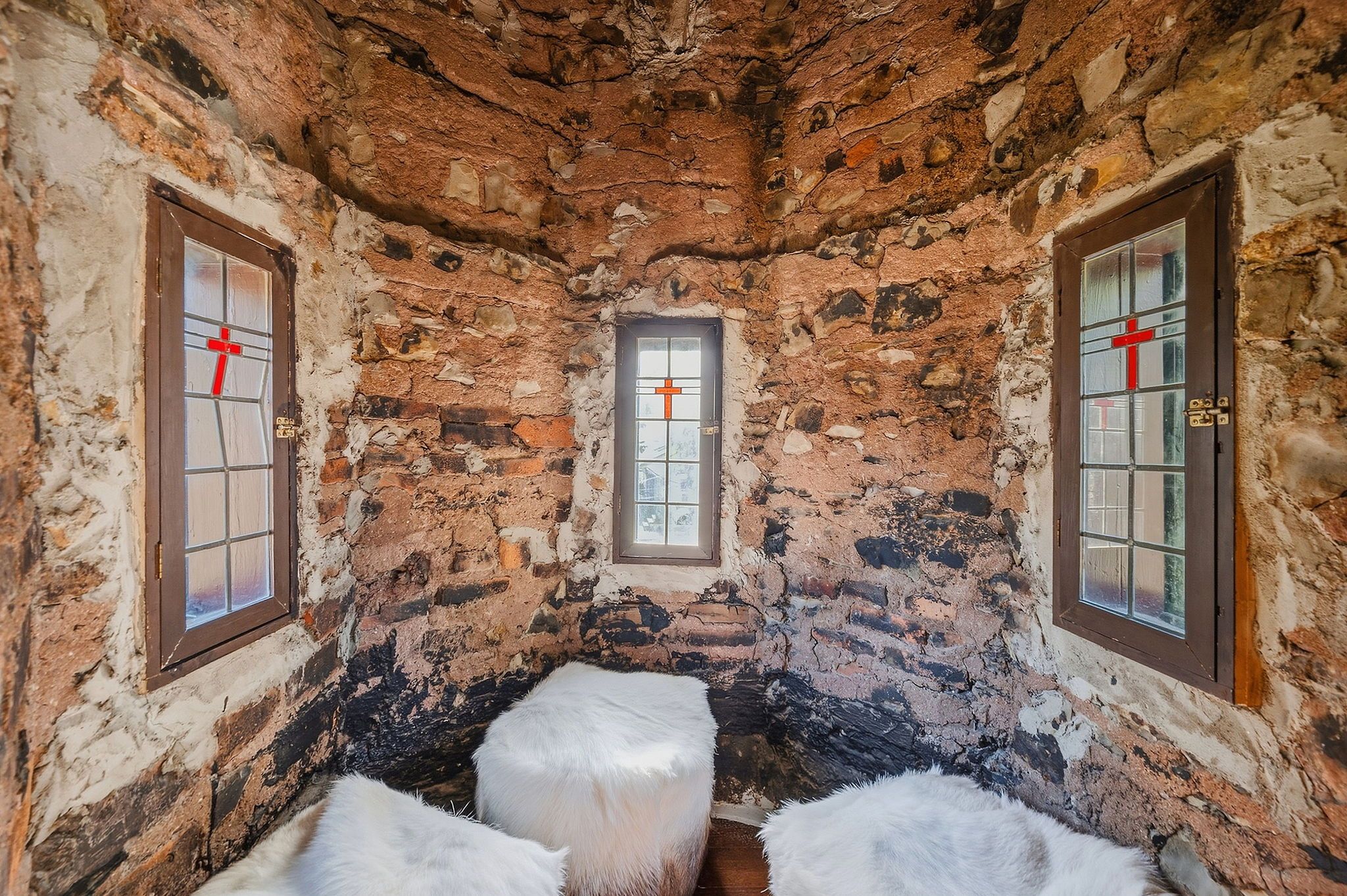 Interior with rough, reddish-brown stone walls and three brown-framed windows; panes show red cross designs. The floor is covered in white fluffy fur rugs, creating a cave-like cozy space.