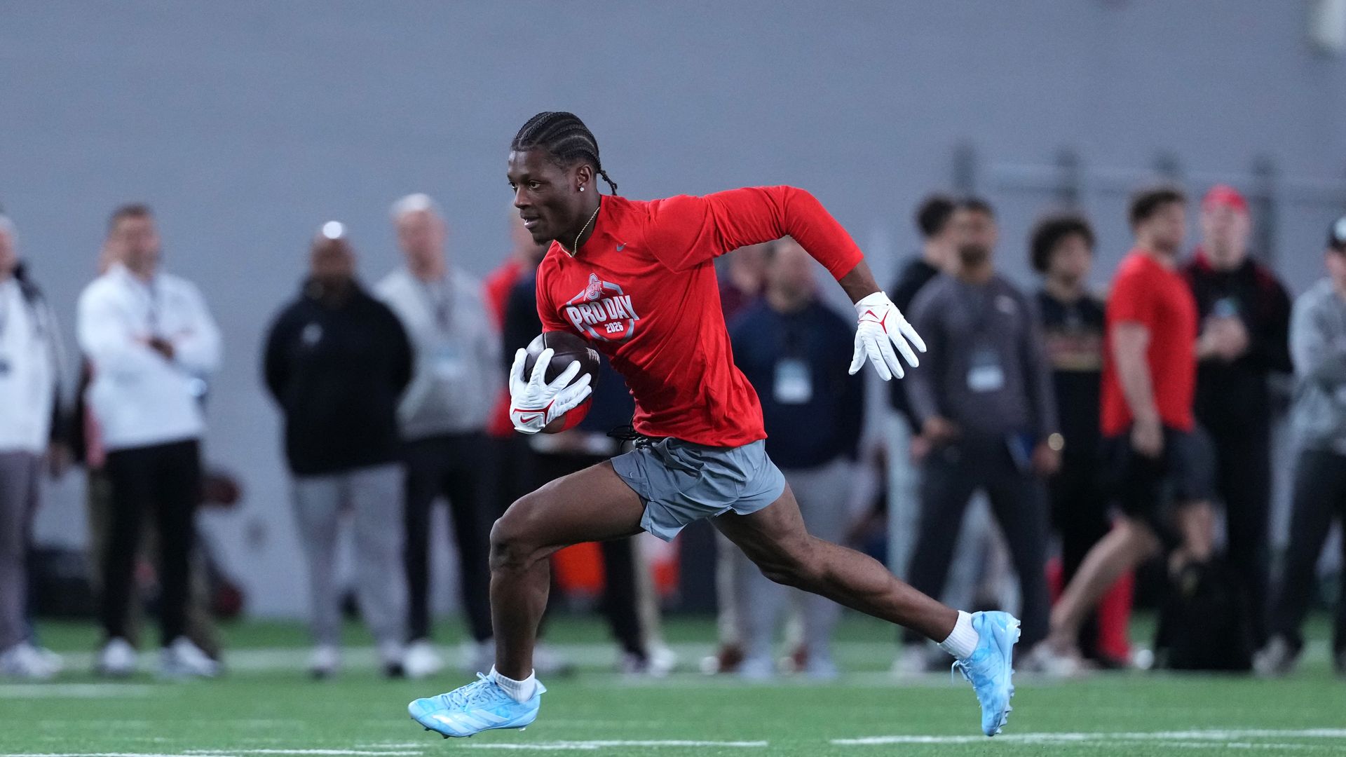 A football player in a red shirt and gray shorts sprints with the ball during a Pro Day drill, wearing white gloves on green turf with a blurred crowd in the background.