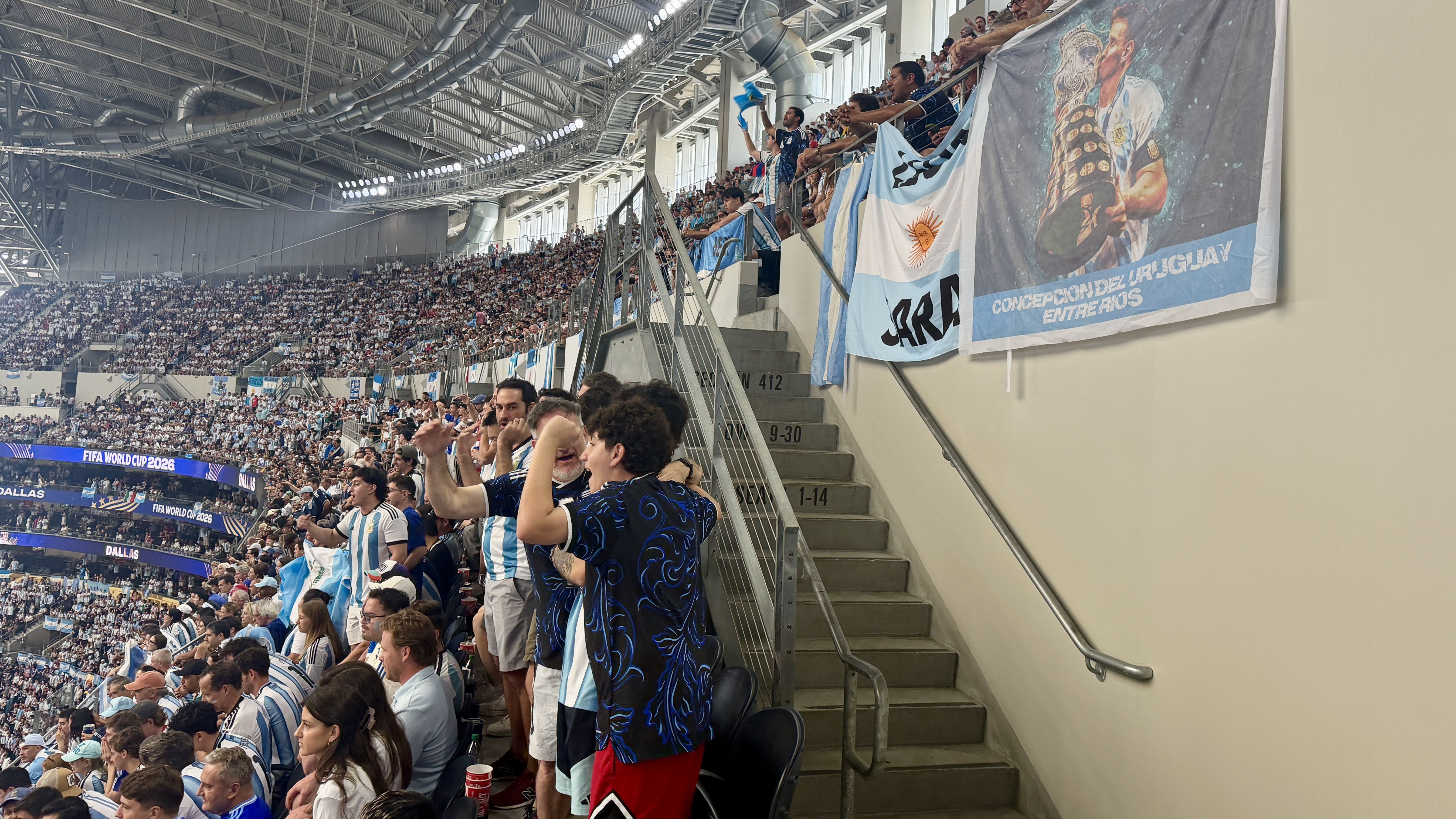 Packed stadium crowd in Argentina colors, fans waving flags. A large banner shows a trophy-bearing player; signage reads "FIFA World Cup 2026", with steel roof and railings overhead.