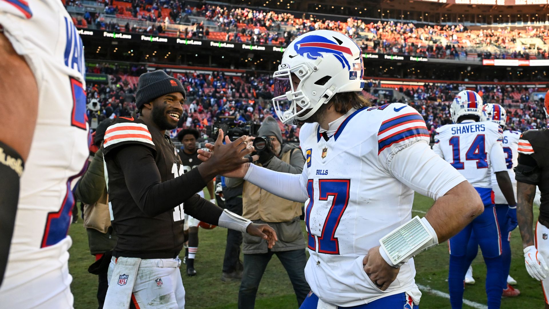 Buffalo Bills players shake hands on the sideline after a game; foreground shows a white-jersey player in a blue-red helmet and a dark-clad teammate, with crowded stands in the background.