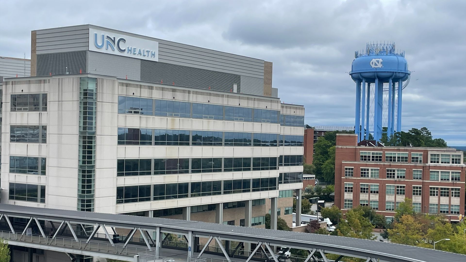 Exterior view of a modern hospital complex with a large UNC Health sign on a gray building; a bright blue water tower with the UNC logo rises in the background under a cloudy sky.