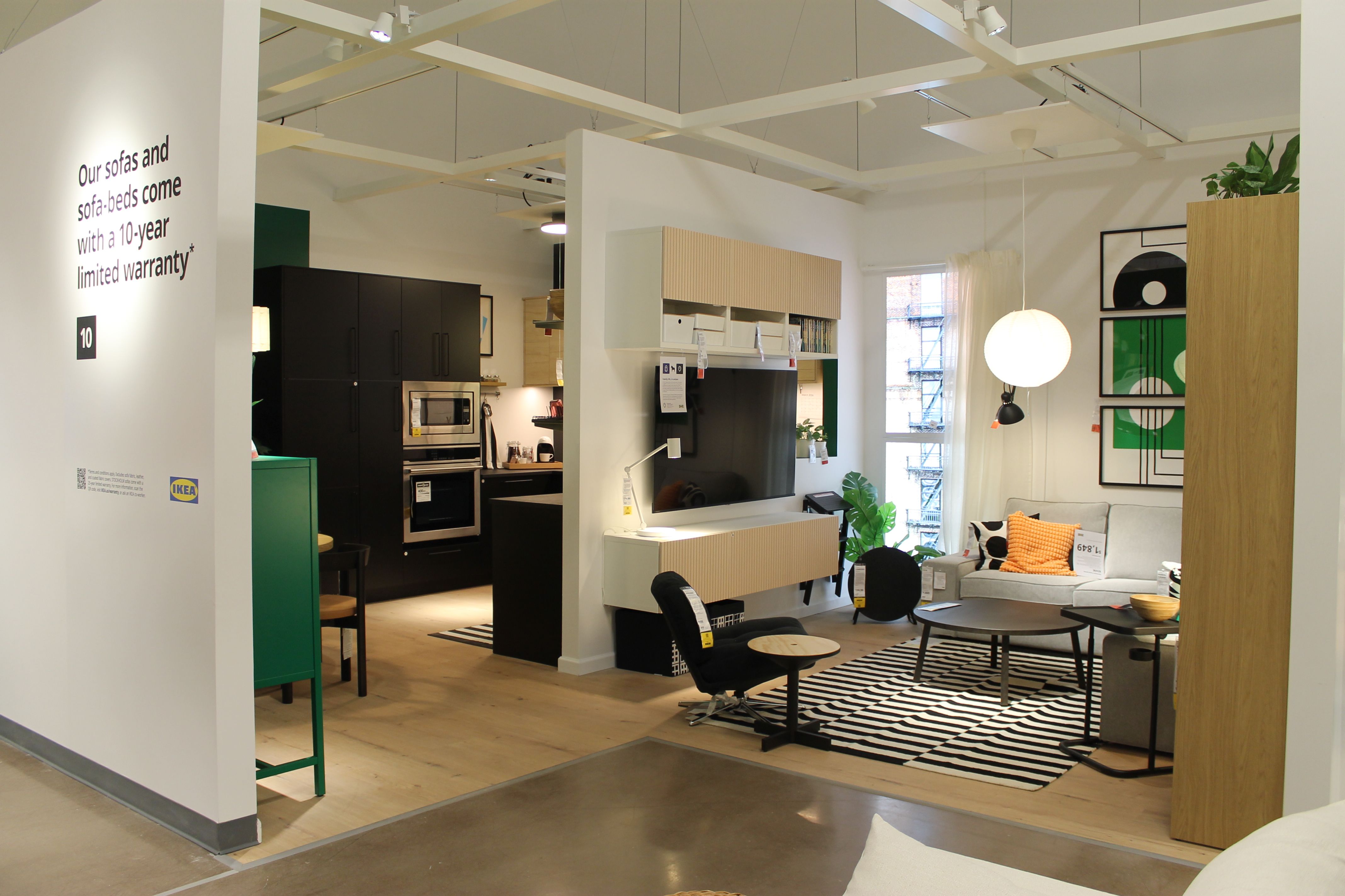 Modern living room and kitchen showroom with black and beige furniture, a striped rug, orange and black pillows, round coffee tables, hanging round lamp, and text on wall about sofas' 10-year limited warranty.