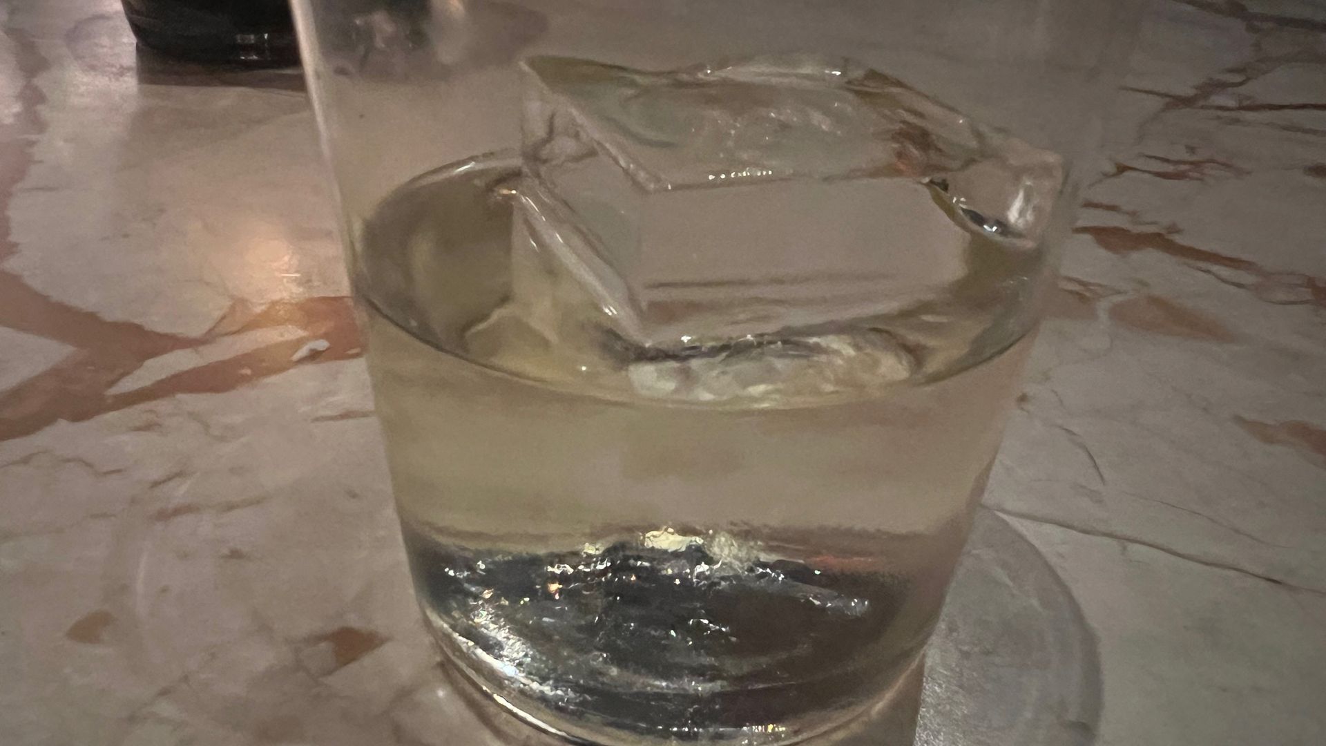 Cocktail glass with large ice cube and clear liquid on a natural stone table lit by candlelight.