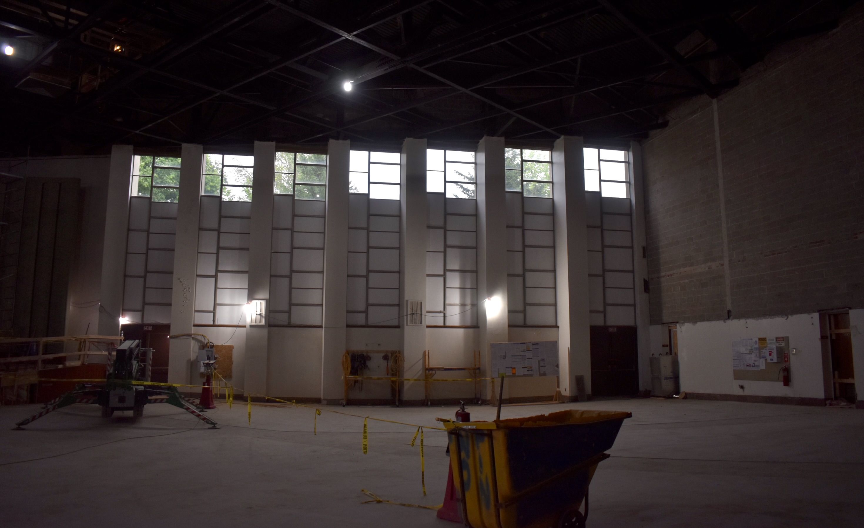 Dim, spacious industrial interior with exposed metal trusses, tall grid windows along far wall, construction equipment, yellow caution tape, and a blue-yellow dumpster in the foreground.