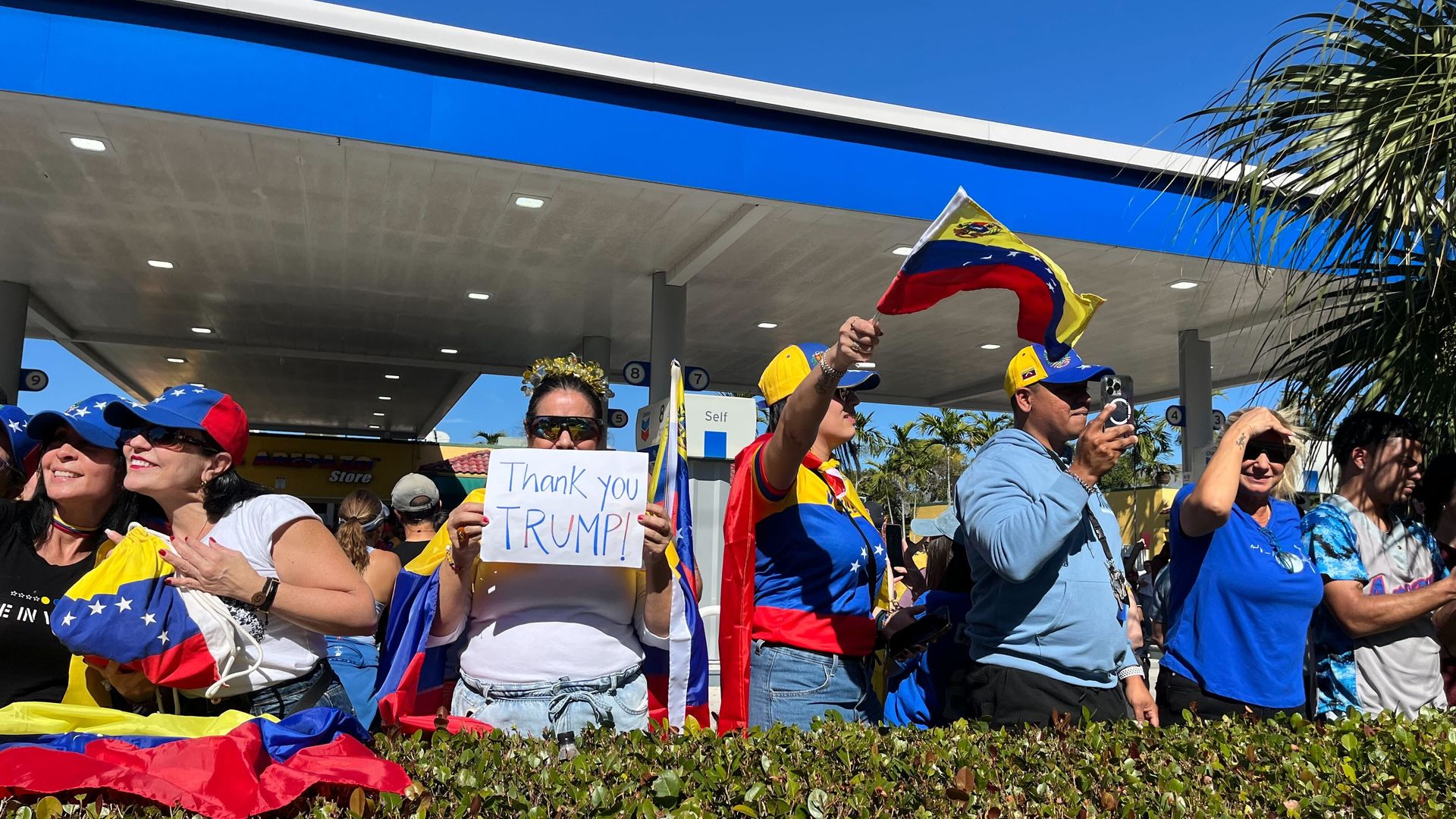Group of people at a gas station with Venezuelan flags and colors, one woman holding a sign that says "Thank you TRUMP!", others waving flags, and a bright blue sky overhead.