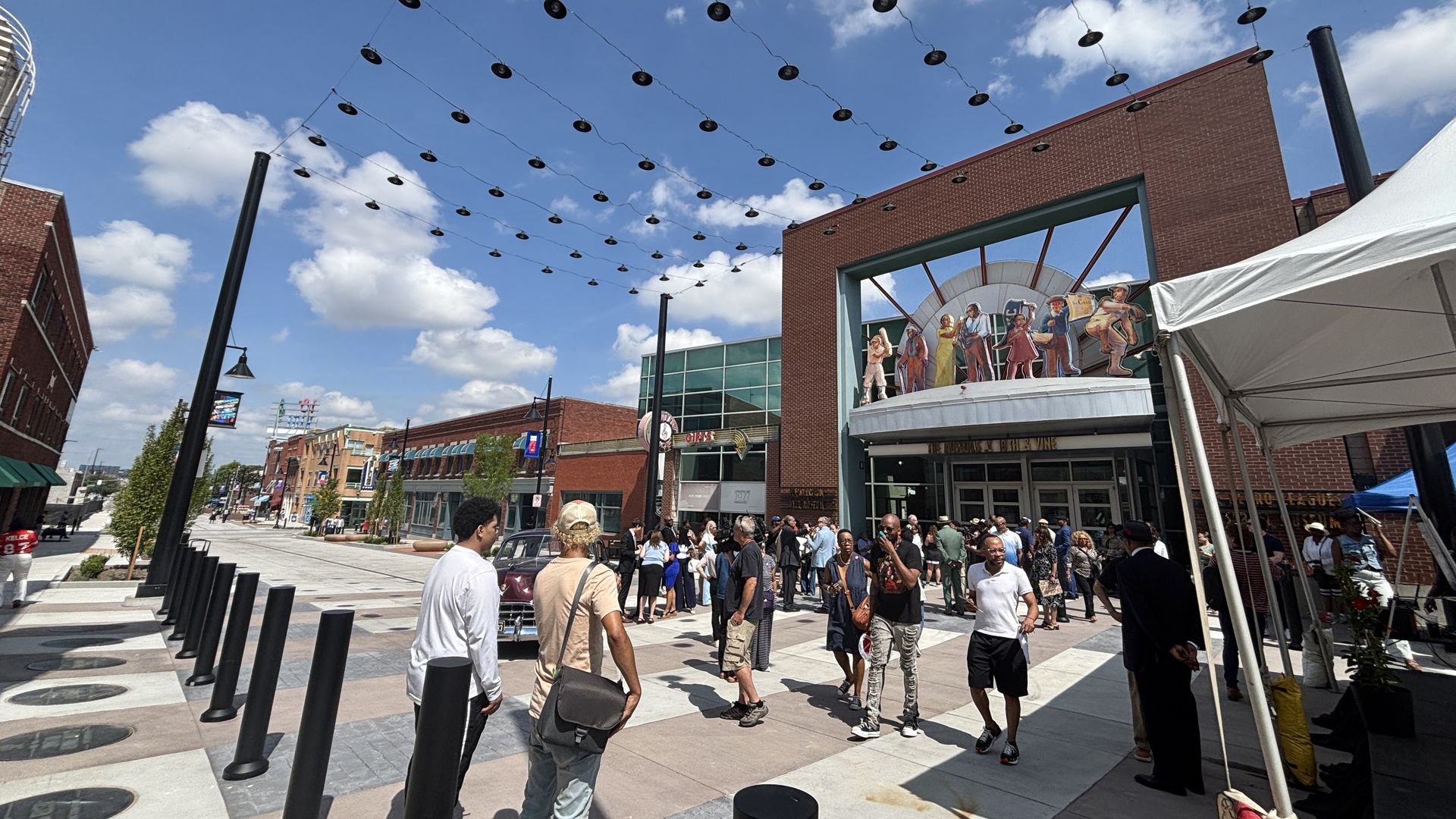 Busy outdoor street scene with string lights overhead, a white event tent on the right, a crowd of people near a brick storefront with a large cutout mural of cartoon figures, blue sky with clouds.