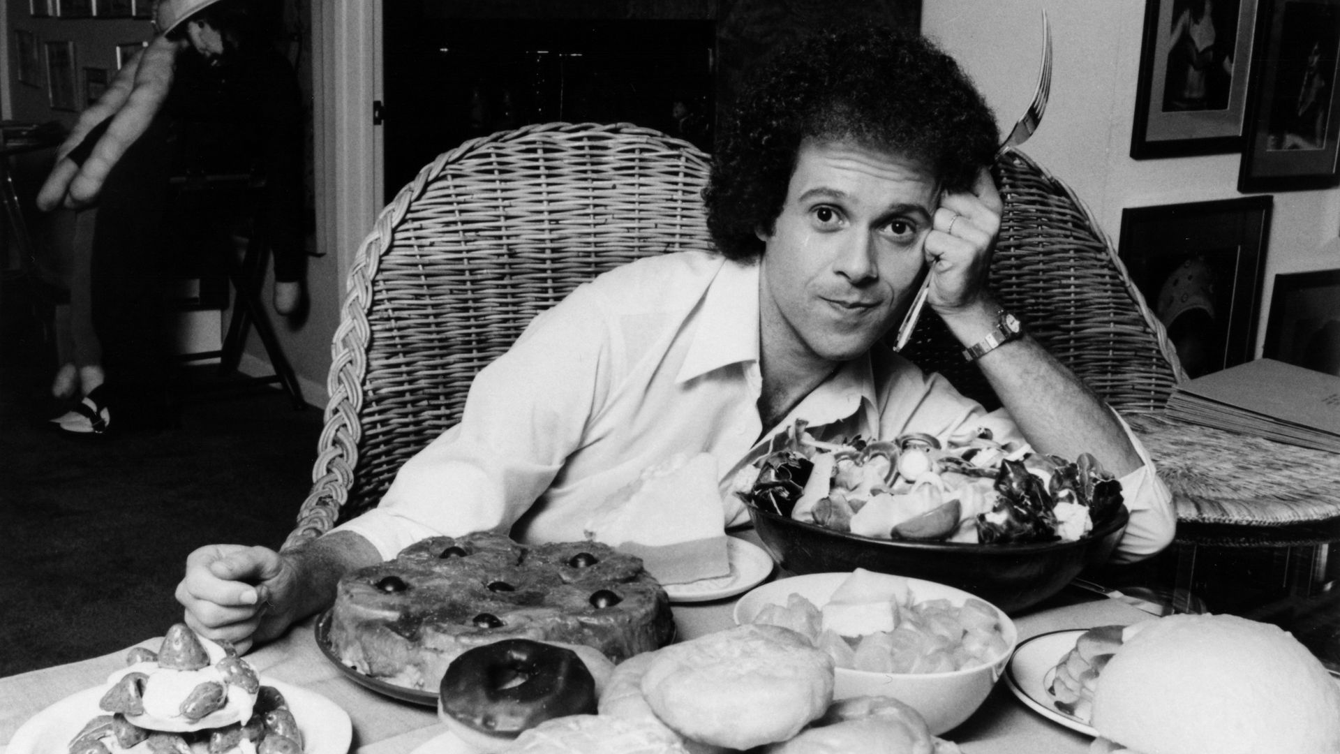 Richard Simmons leans on his elbow on a table filled with food in an archival black and white photo.