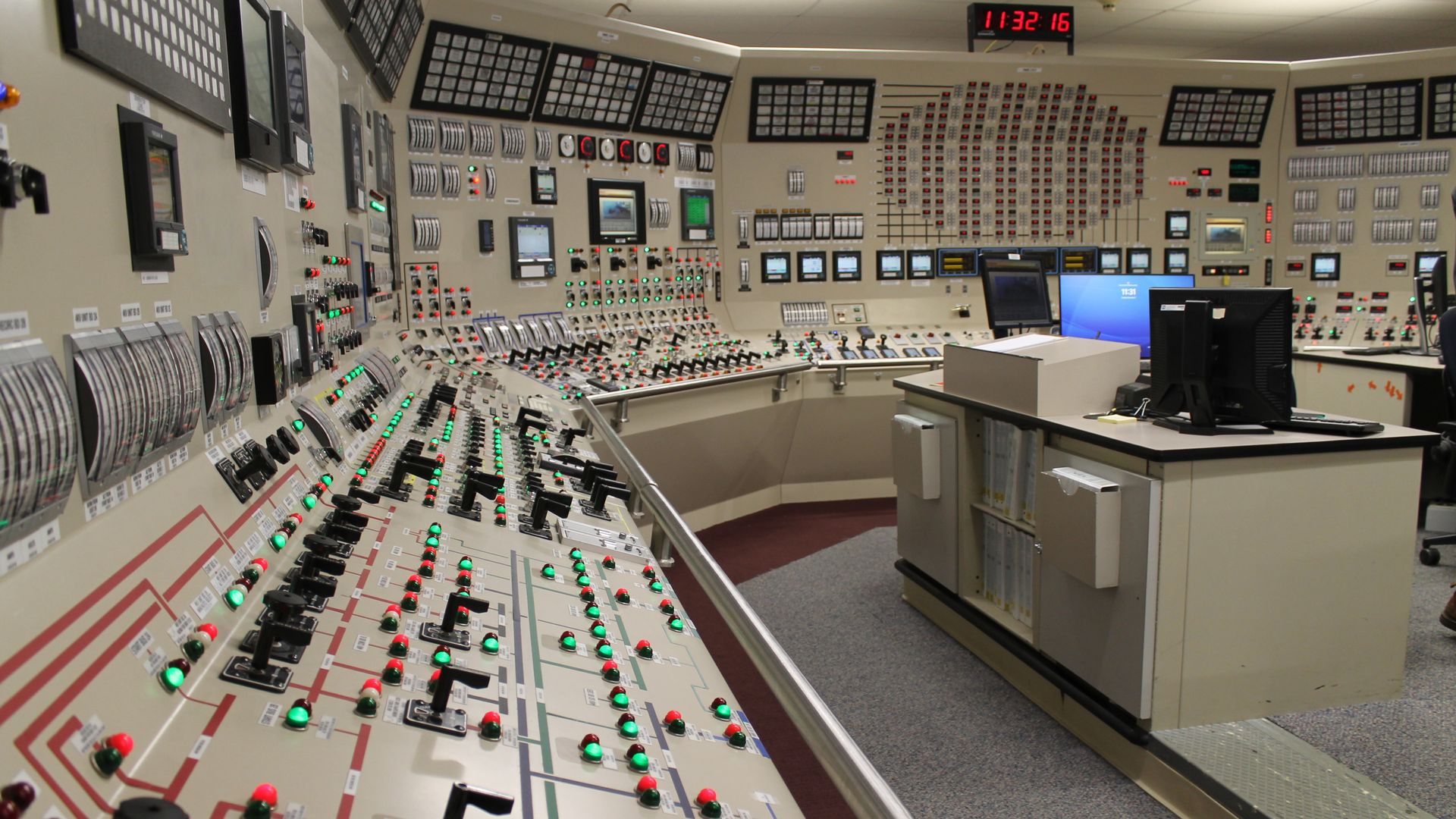 Wide shot of a vintage control room with beige panels, rows of switches and green/red indicator lights, dials, and several monitors; a desk with a computer on the right.