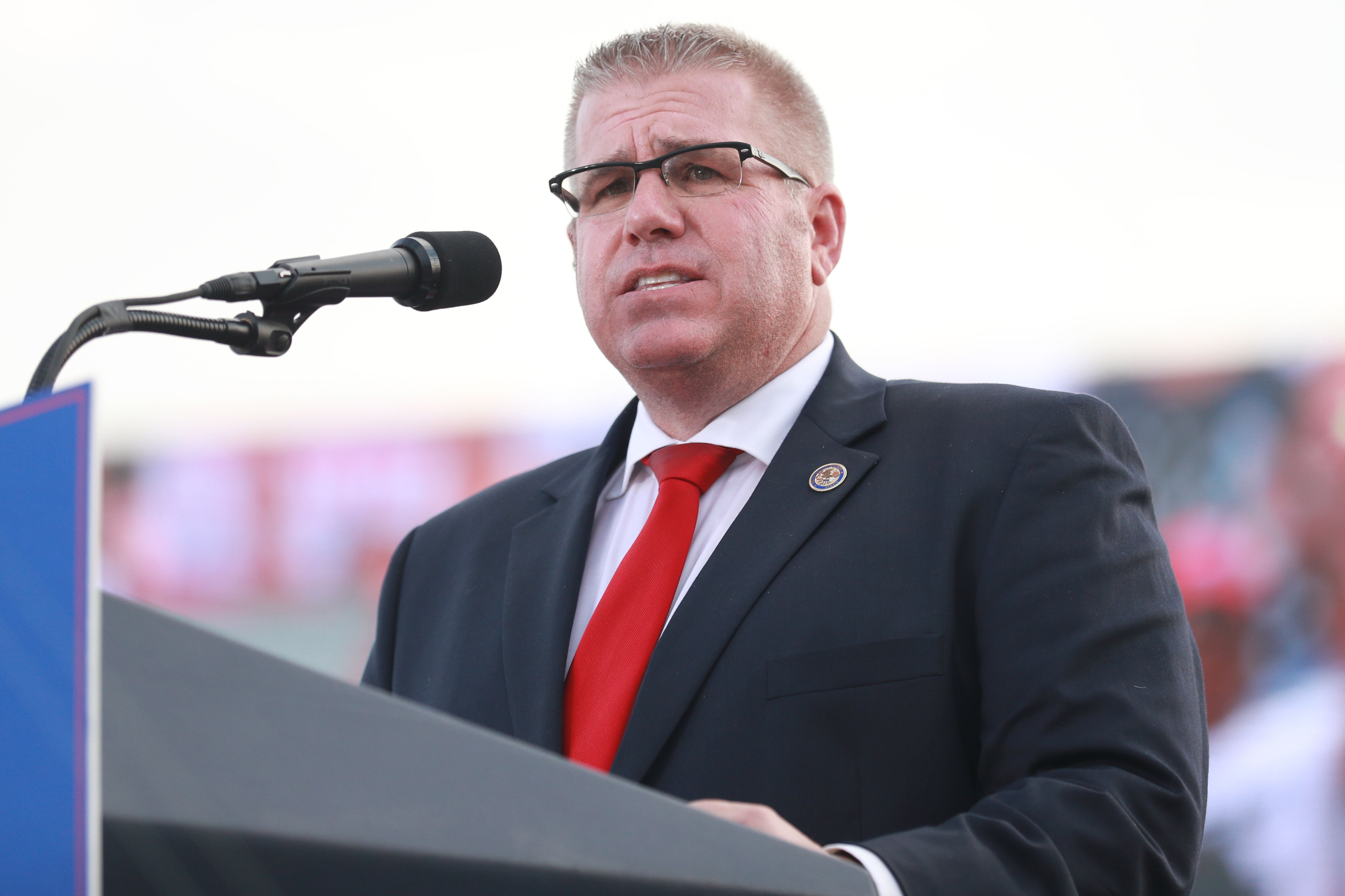 Man wearing glasses, a black suit, white shirt, and red tie speaking at a podium with microphone, light background, and blurred crowd behind.
