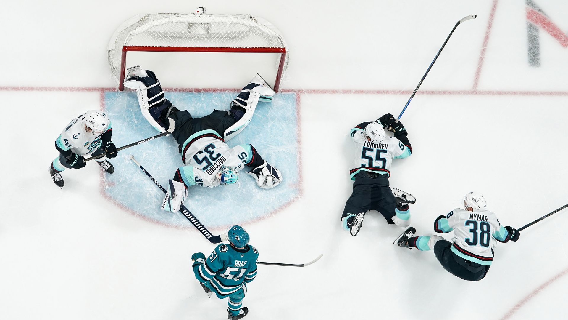 An overhead view of  a hockey net with players sprawled around it. 