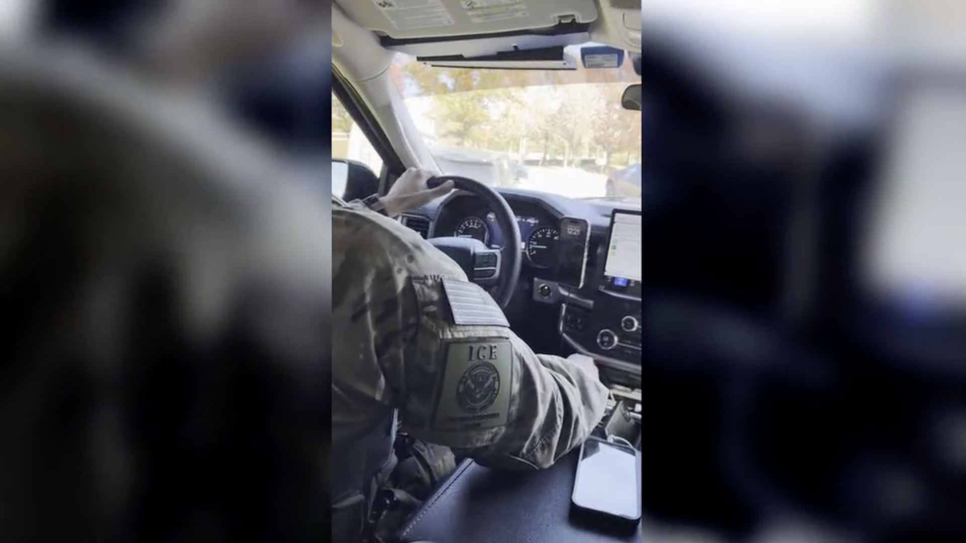 View from inside a vehicle showing a person in camouflage uniform with an ICE patch on sleeve driving. The dashboard and smartphone are visible with a bright outdoor scene beyond the windshield.