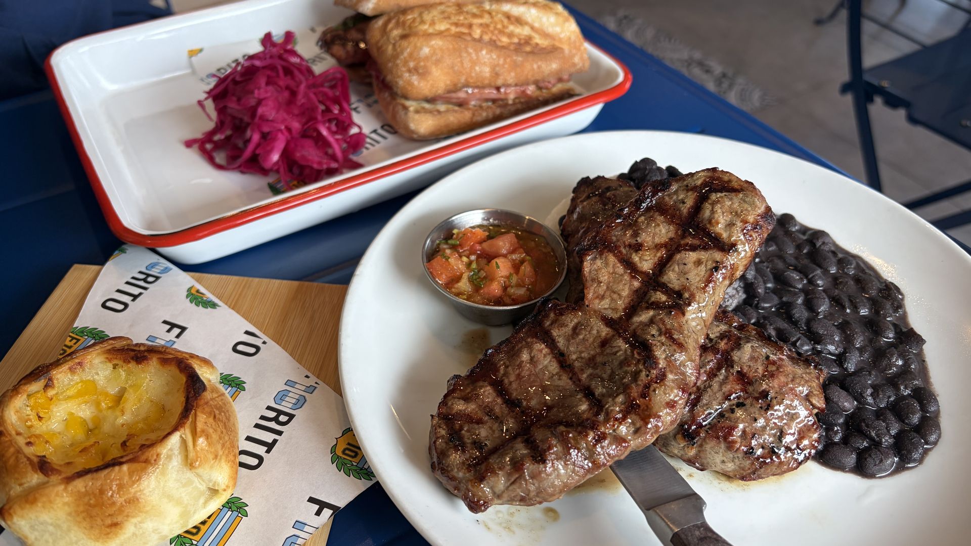 Plate with grilled steak, black beans, and salsa; sandwich with pickled red cabbage on a tray; bread roll with melted cheese on a wooden board, all on a blue table.